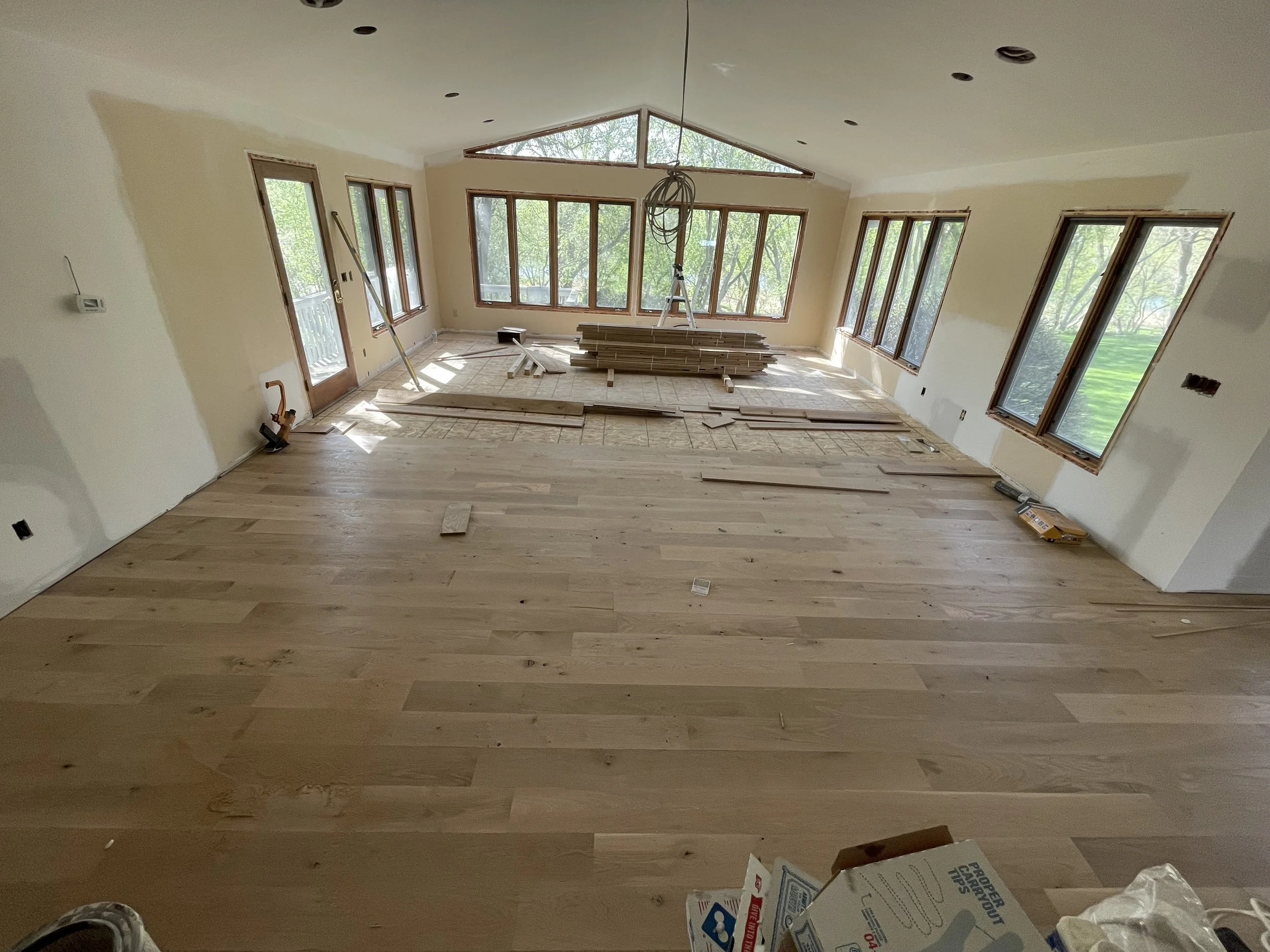 Room under renovation with installed hardwood flooring and unfinished walls, large windows, and construction materials scattered across the floor.