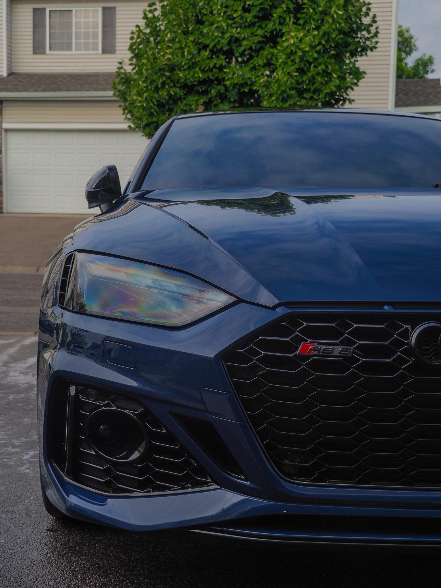 Front view of a blue Audi RS5 parked in a residential driveway, focus on grille and headlights, house and tree in background.