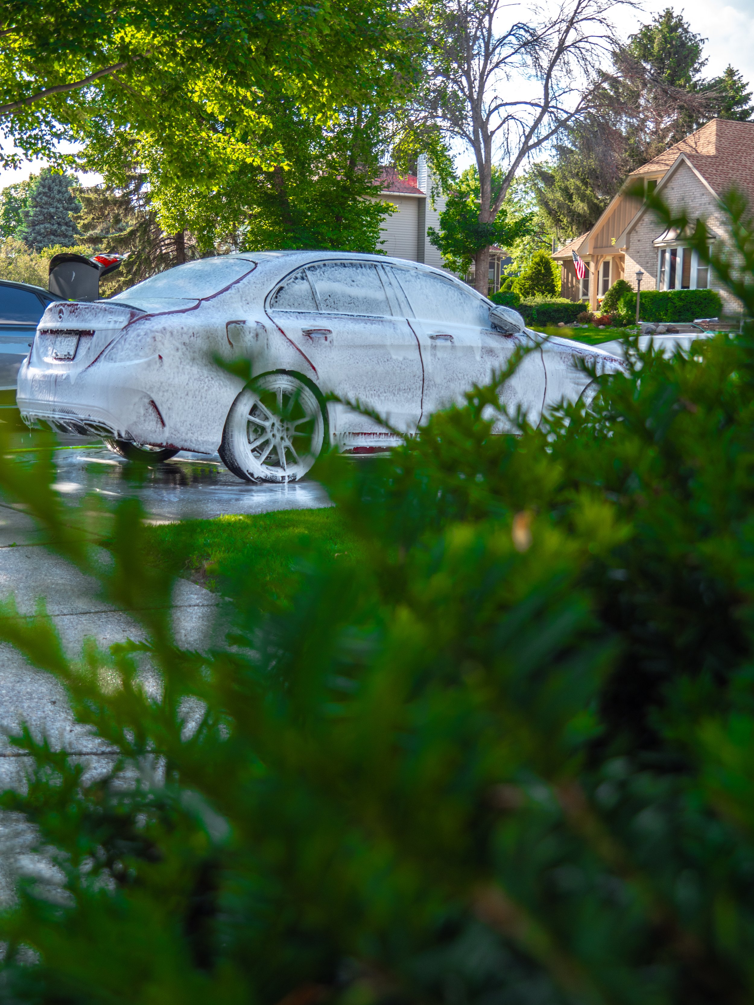 Car covered in soap for washing, parked on a driveway, surrounded by greenery and residential houses.