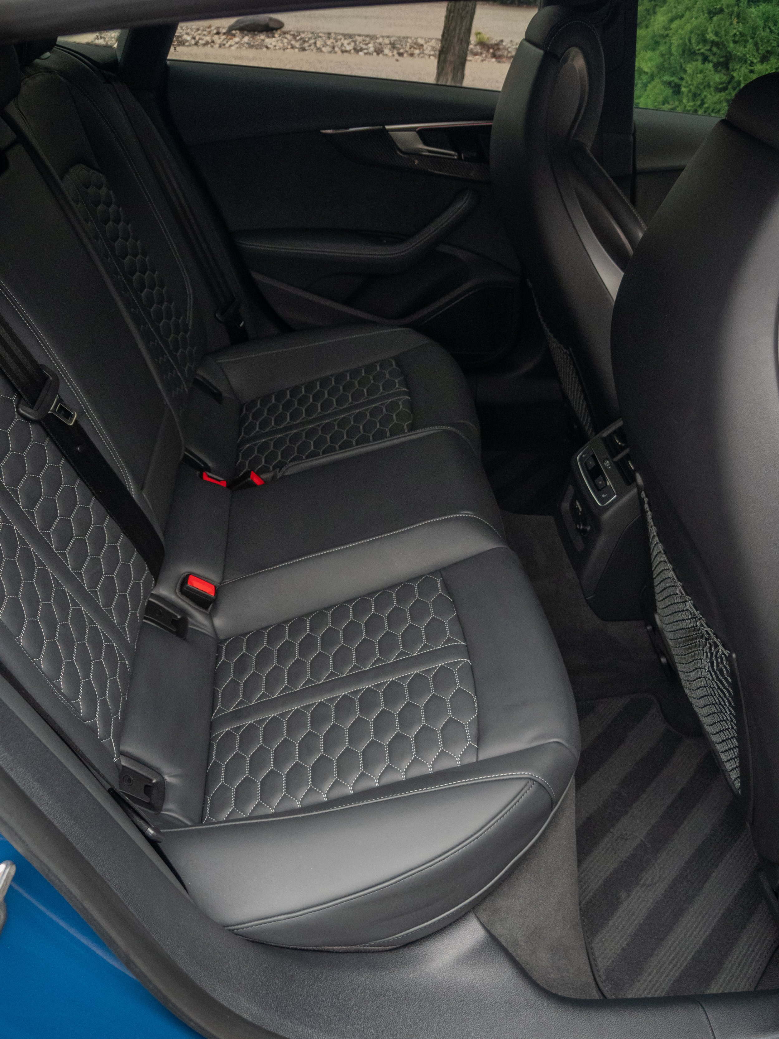 Backseat interior of a car with black leather seats featuring hexagonal stitching, visible car door and carpeted floor mat.
