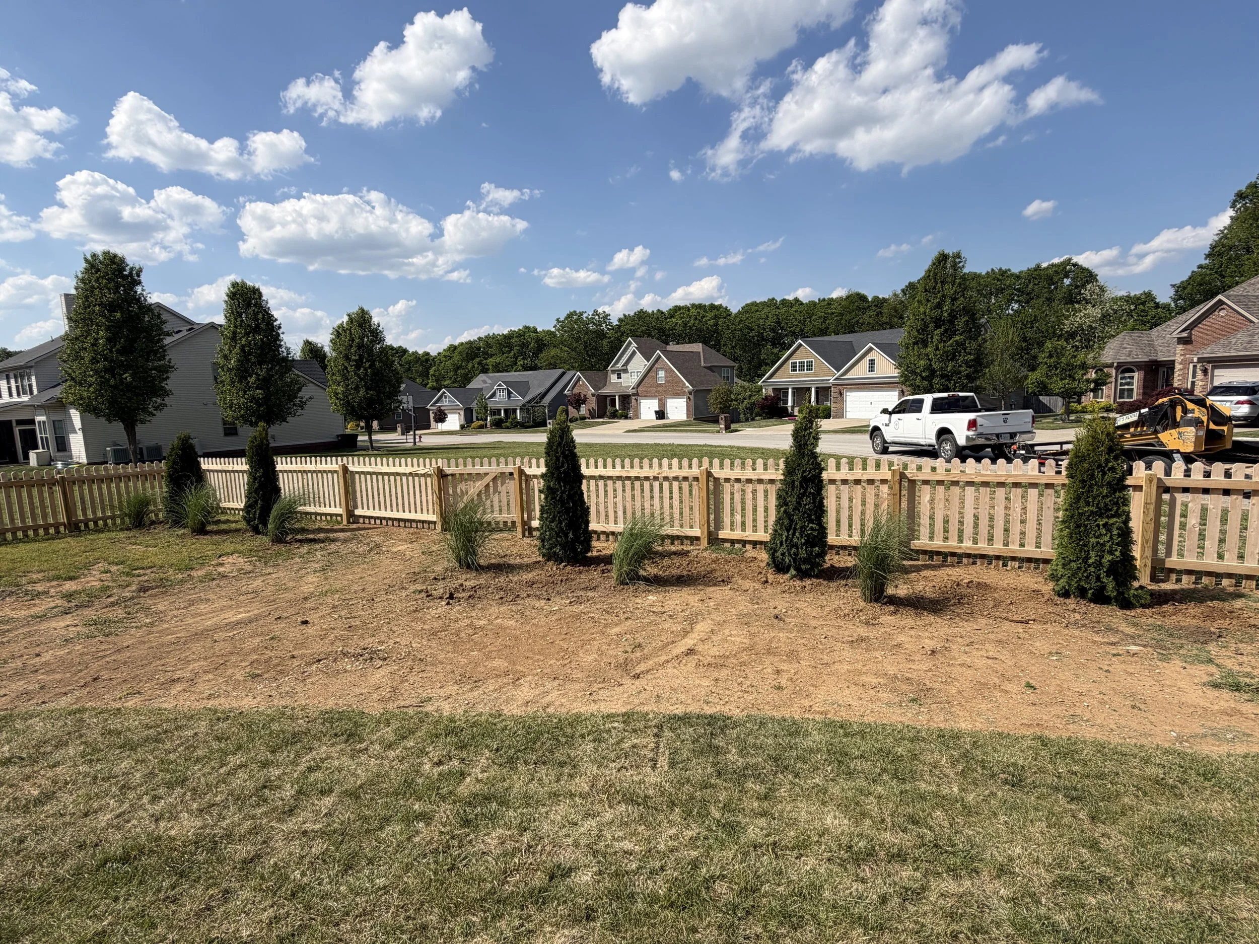 Suburban neighborhood with new homes, wooden fence, landscaping, parked pickup truck, and sunny blue sky with clouds