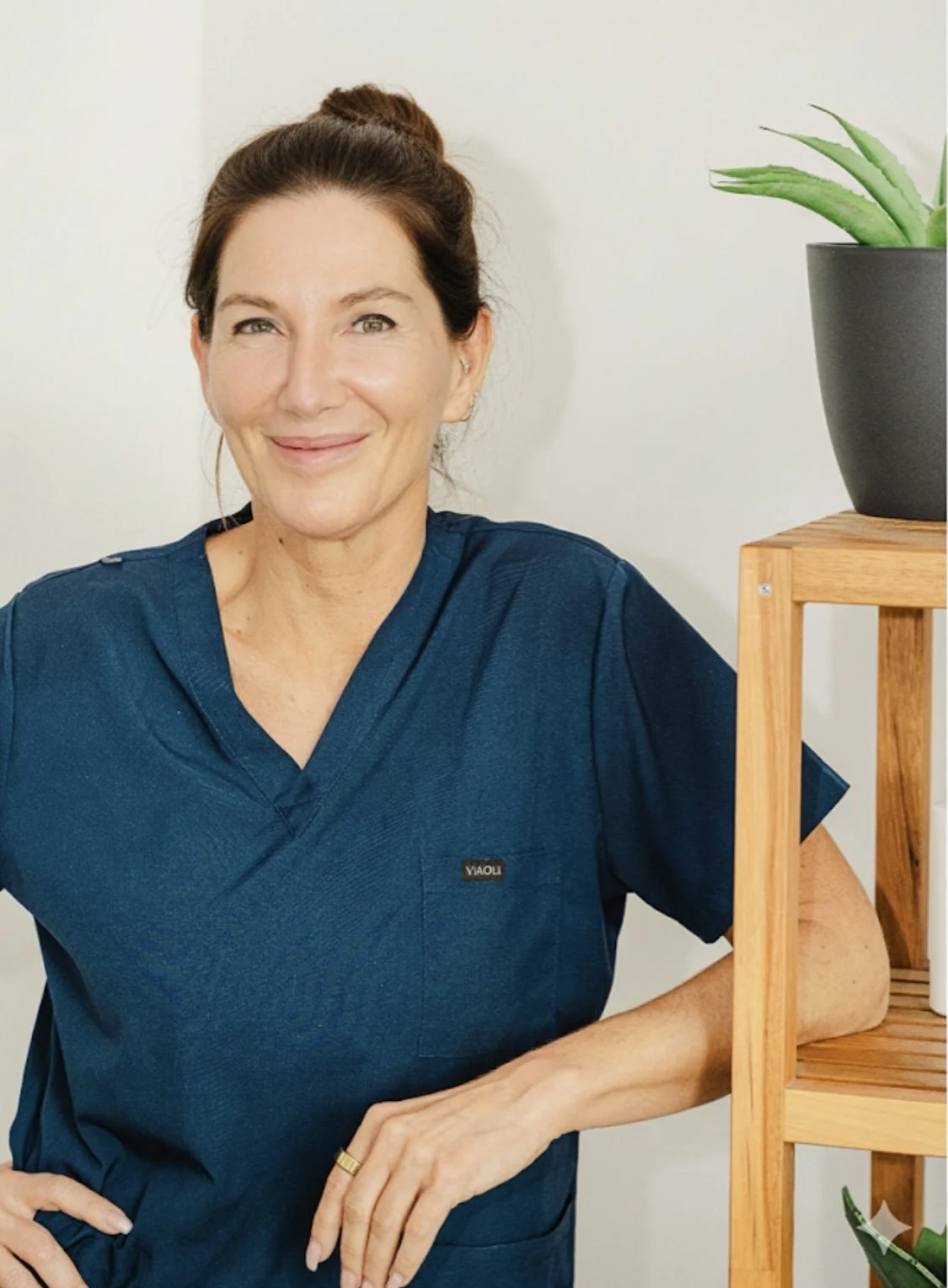 Frau Annette Roscher in blauer medizinischer Kleidung steht neben einem Holzregal mit einer grünen Pflanze in einer schwarzen Vase.