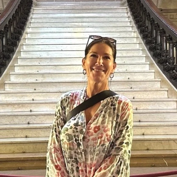 Eine lächelnde Frau steht vor einer großen Treppe in einem öffentlichen Gebäude, trägt eine bunte Bluse, eine Umhängetasche und tragt Brillen auf dem Kopf.