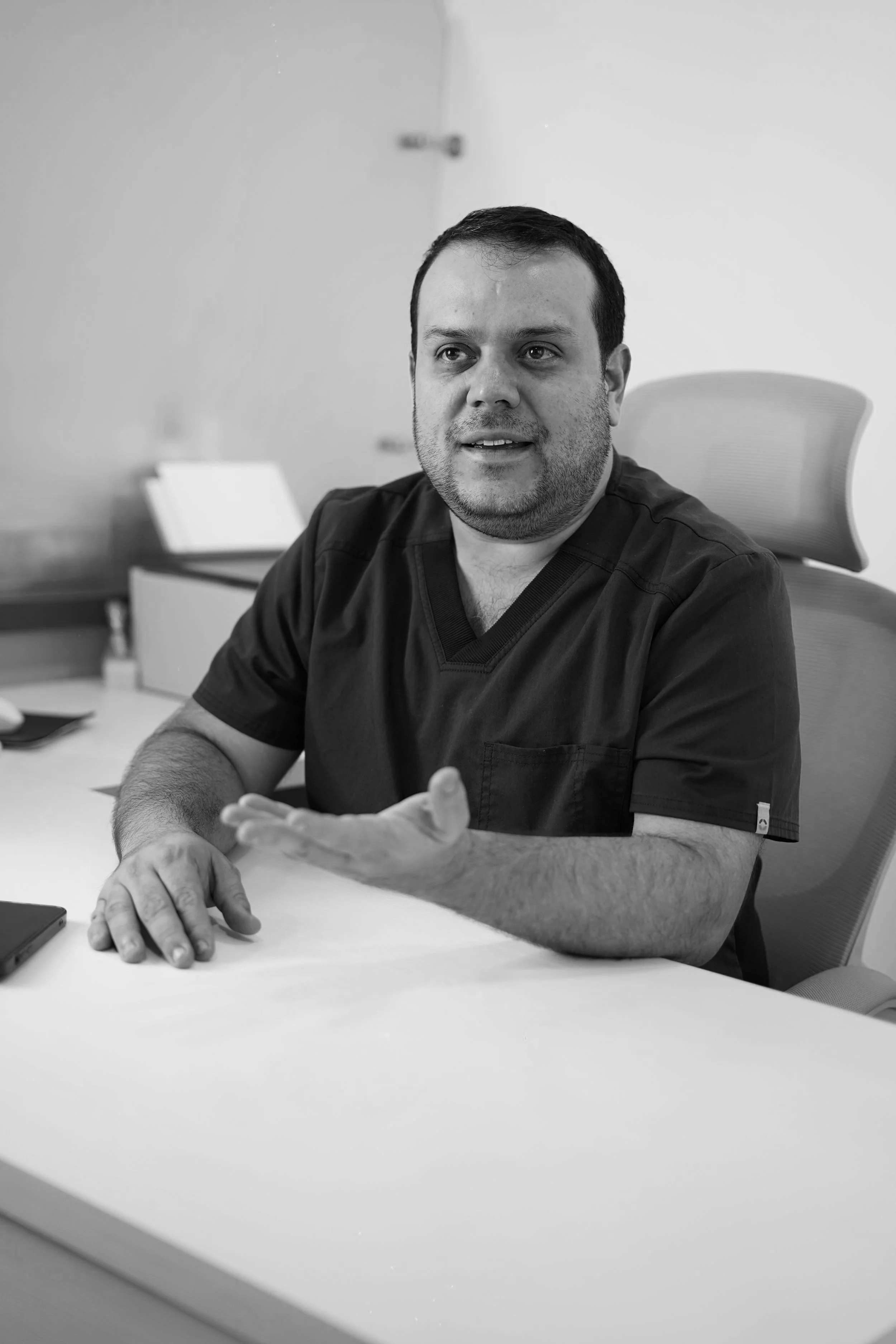 Hombre en una sala de consulta médica, vestido con uniforme negro, sentado en una mesa, en una silla ergonómica, hablando y gesticulando con la mano derecha.