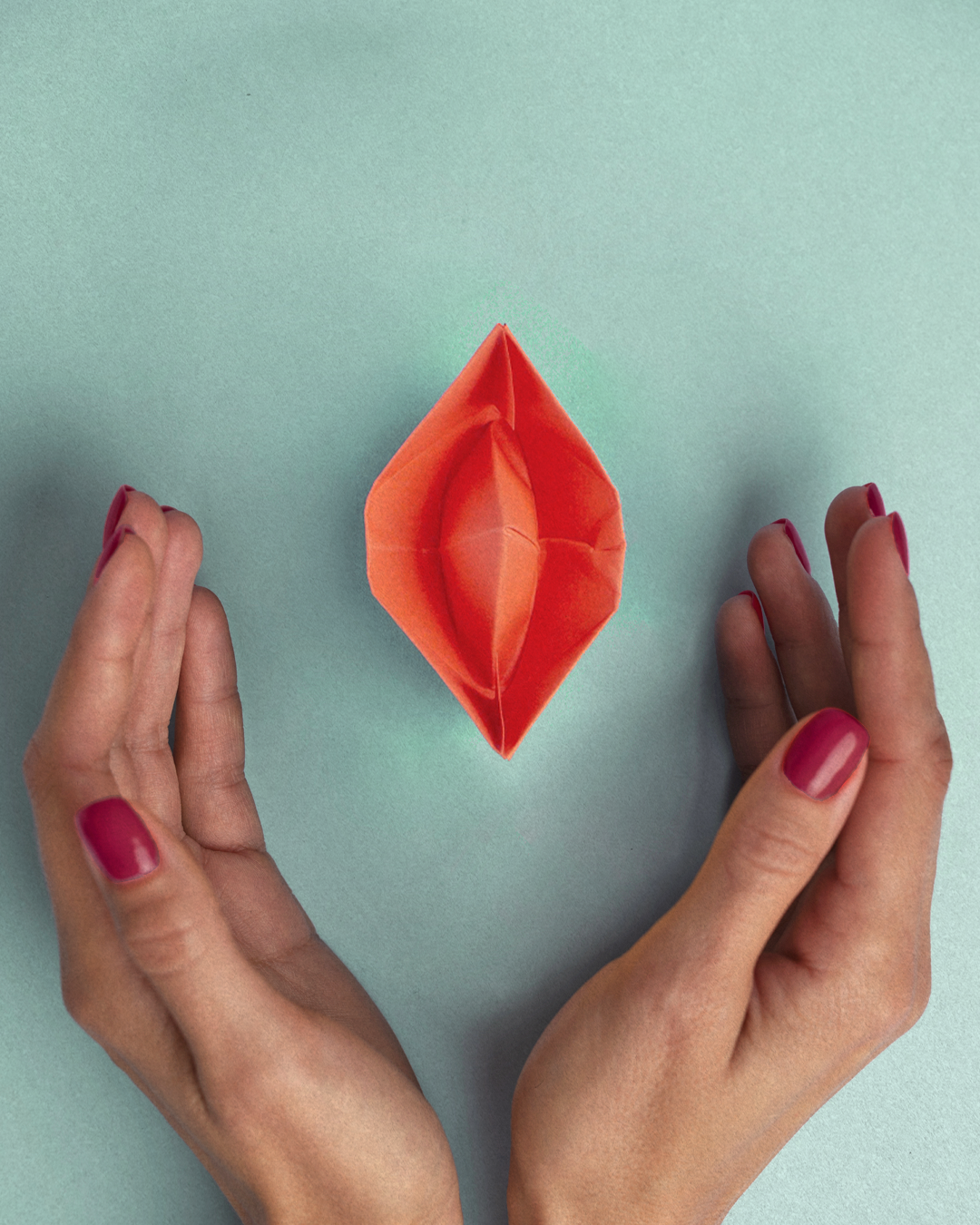 Manos con uñas pintadas sosteniendo un barco de papel naranja sobre fondo azul claro.