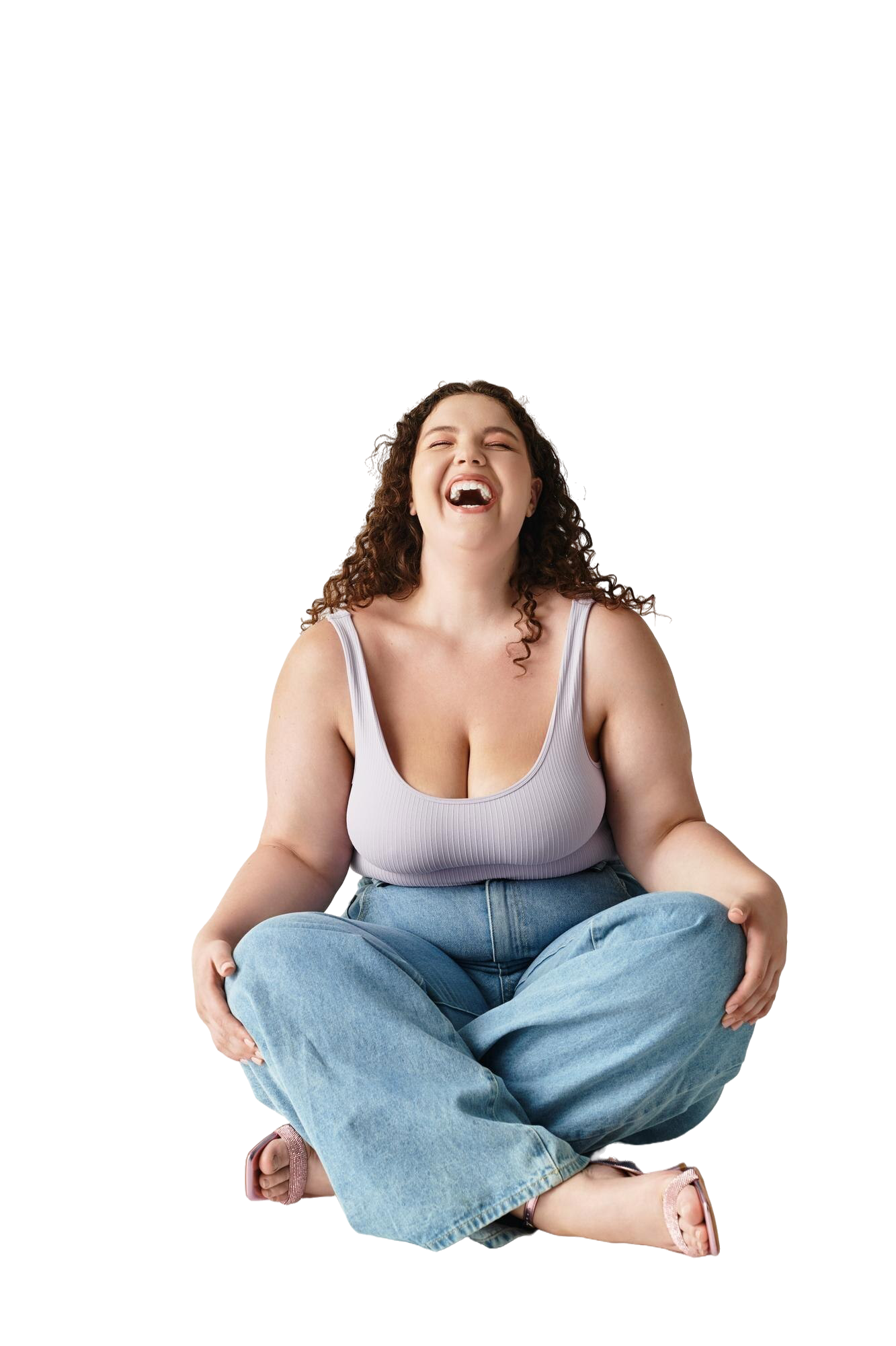 Mujer con cabello rizado, sonriendo y riendo, sentada con las piernas cruzadas, vistiendo camiseta sin mangas gris y pantalones vaqueros de amplio corte.