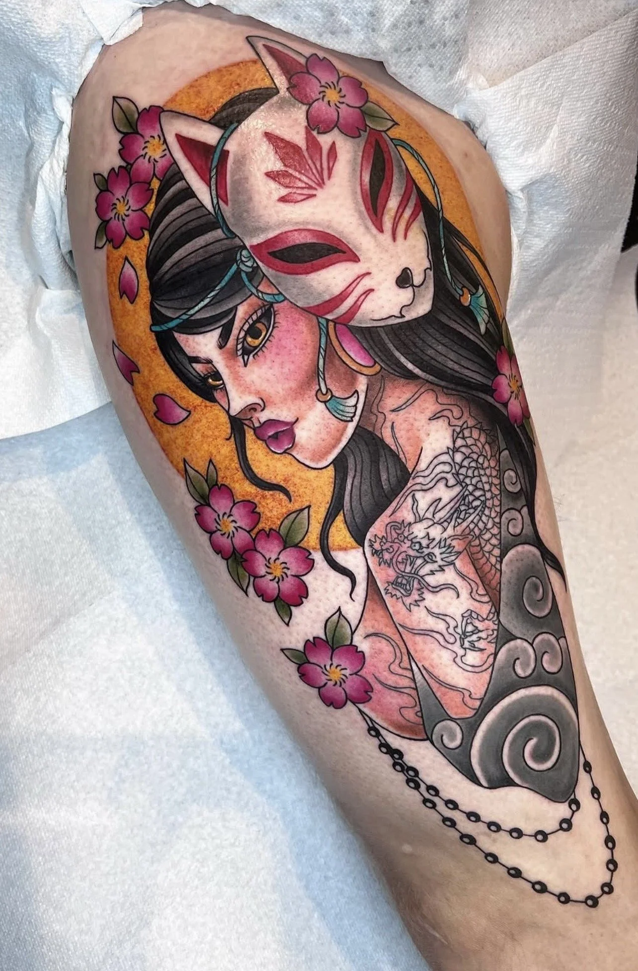 Tattoo of a woman with dark hair and gold earrings, surrounded by pink flowers and an orange background, with a masked fox and a dragon on the arm.