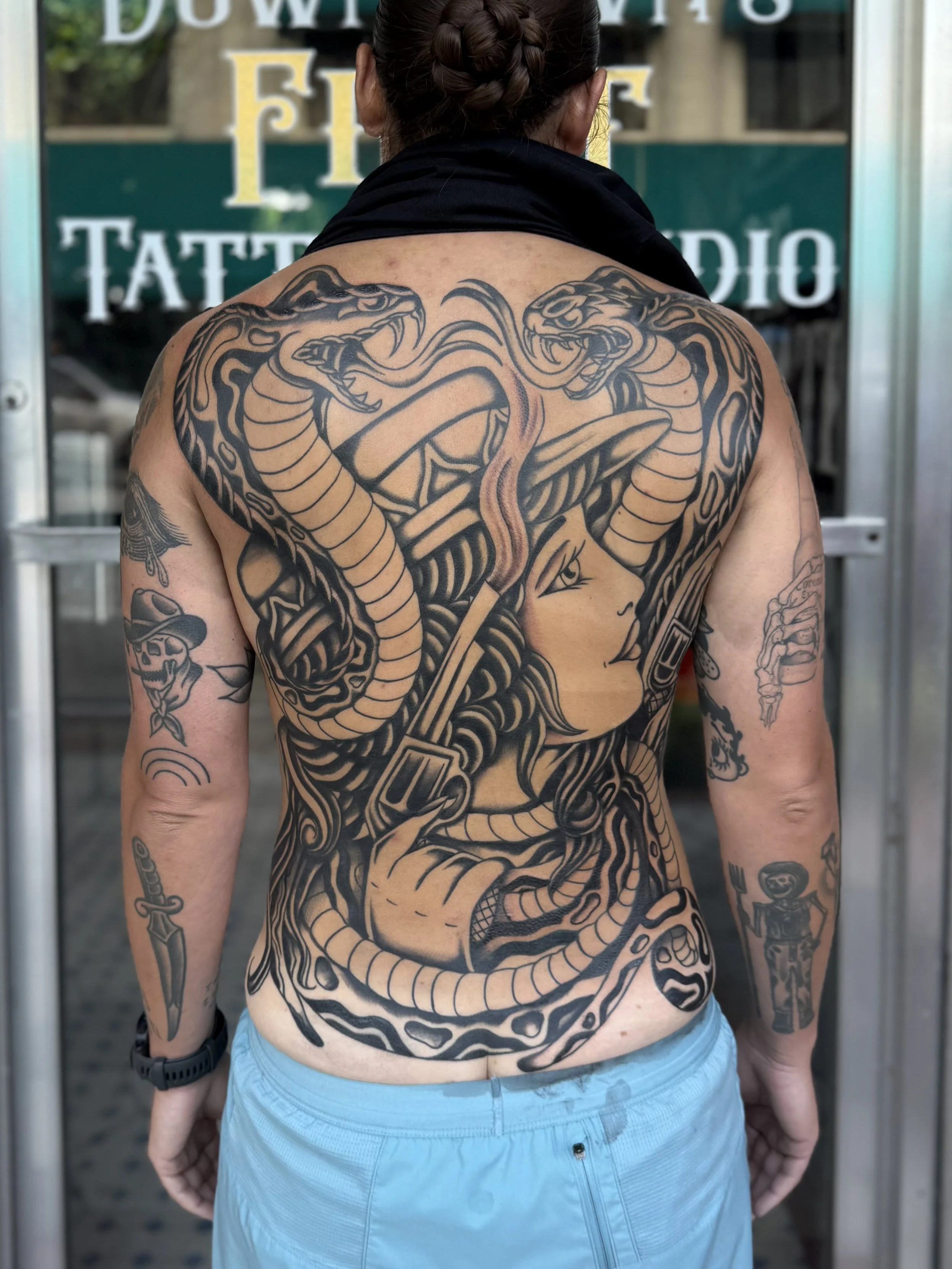 A person with their back facing the camera shows a large intricate tattoo of a snake coiled around a sword on their back. The person has dark hair styled in braids and is wearing a black scarf and light blue shorts.