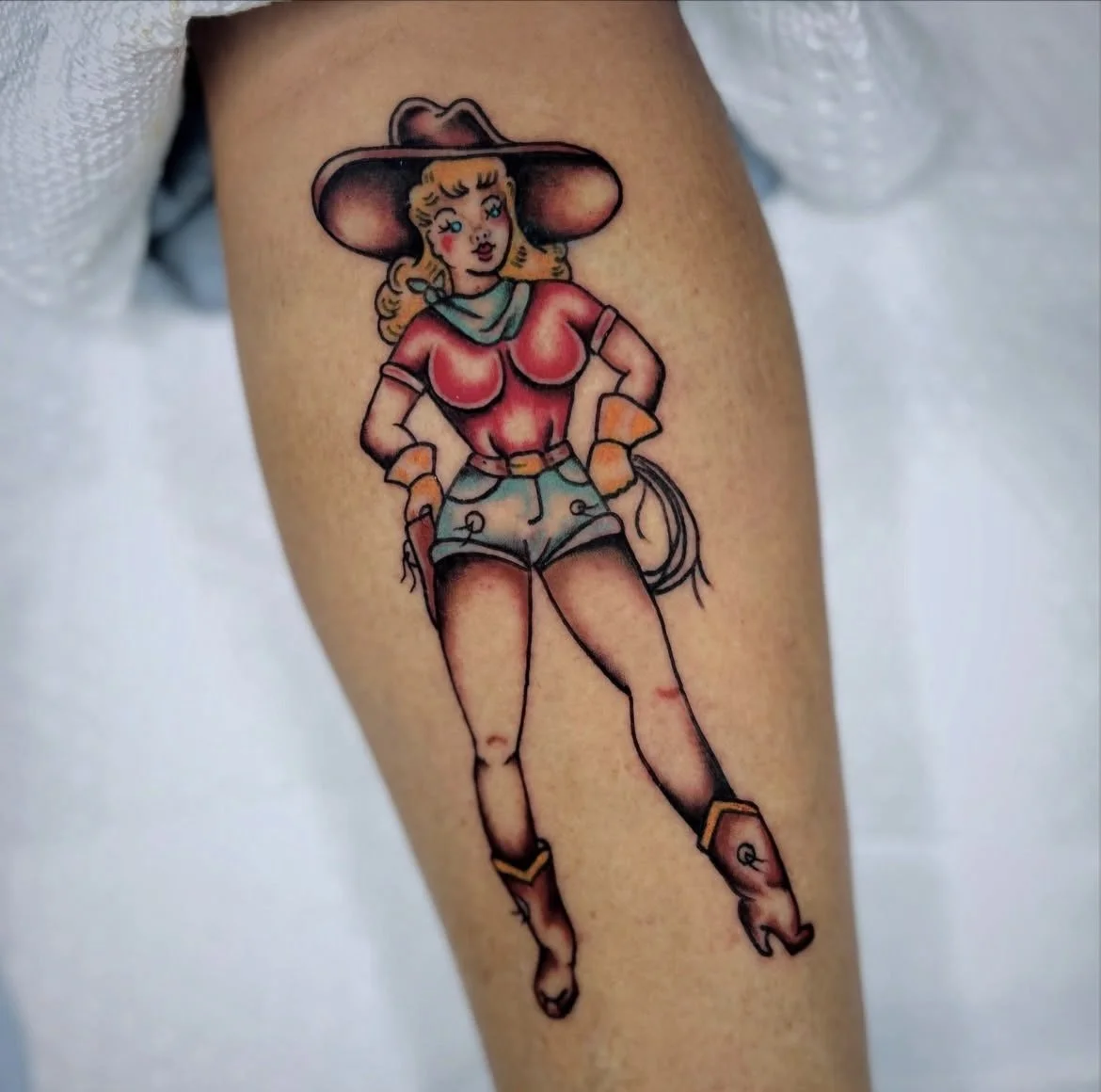 Tattoo of a cartoon-style woman in Western cowboy attire, including a wide-brimmed hat, red shirt, denim shorts, gloves, and boots.