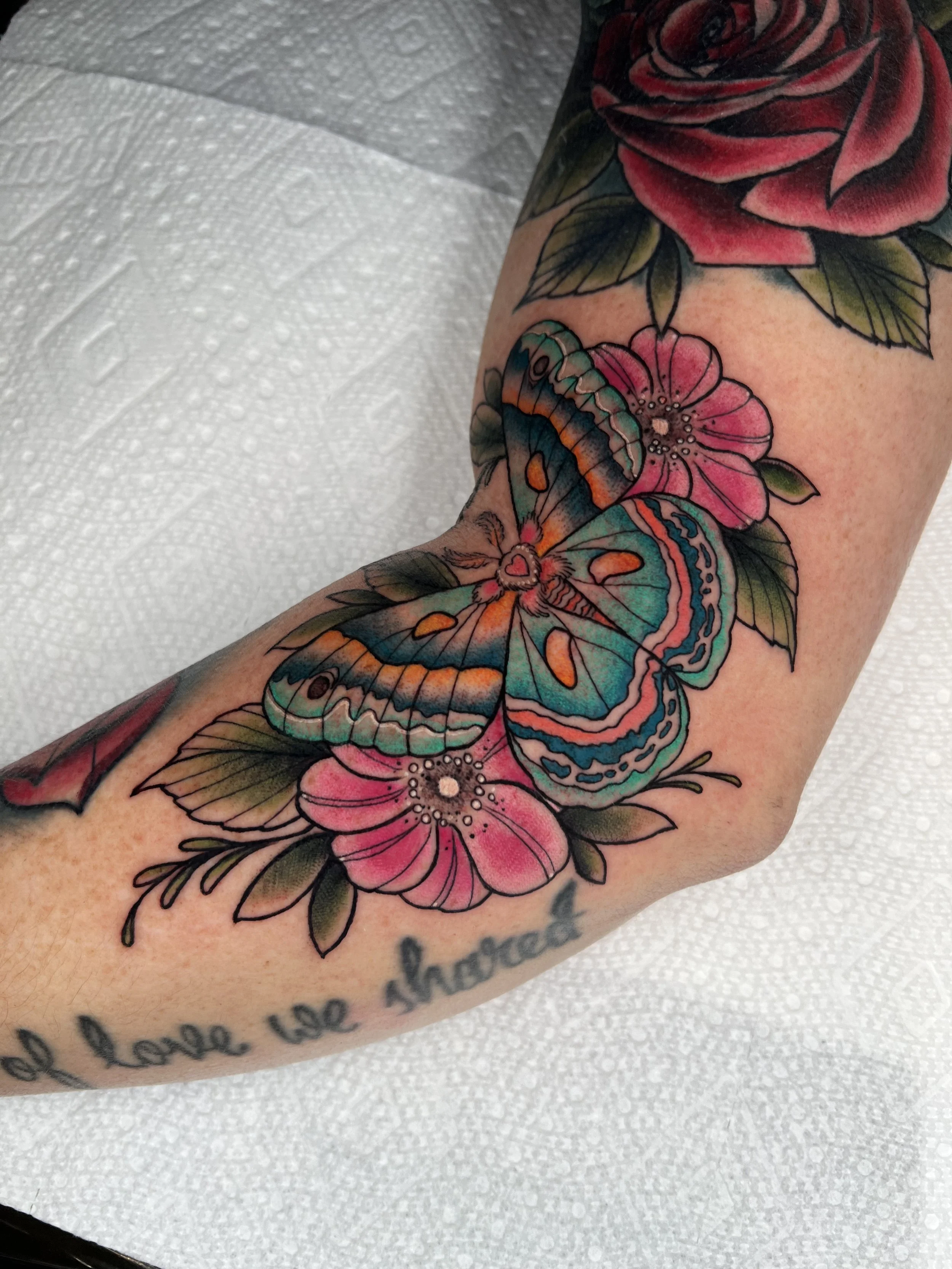 Colorful tattoo of a pink flower, butterfly, and leaves on a person's arm with a script text underneath.