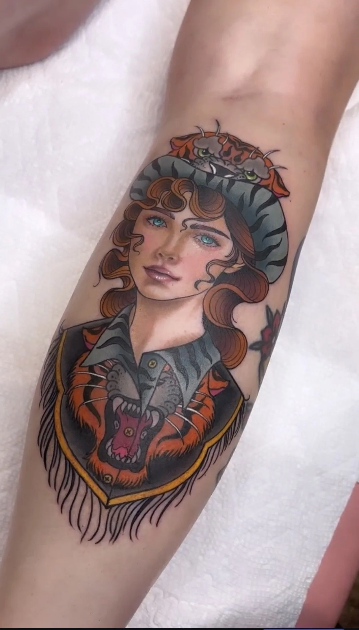 Tattoo of a woman with blue eyes and curly hair, wearing a hat and jacket, with a tiger head tattoo and a shield with a tiger's face on the arm.