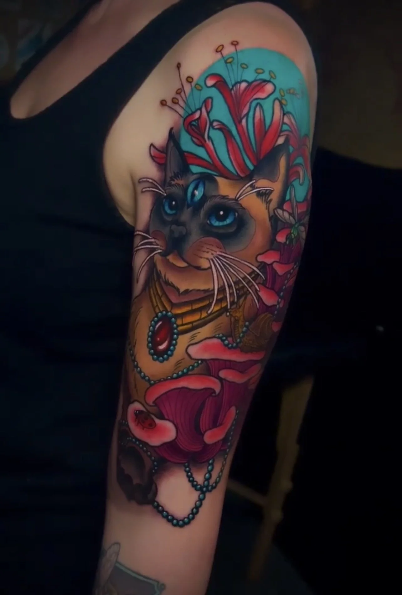 Colorful tattoo of a cat with blue eyes, surrounded by red flowers, a necklace, and a ladybug on the forearm.