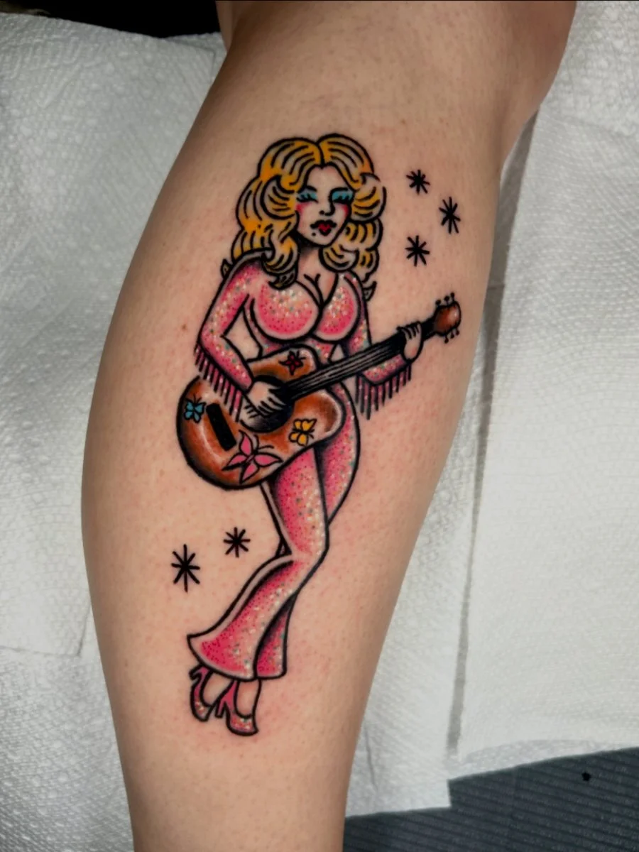 Tattoo of a woman with curly blonde hair in a pink jumpsuit holding a guitar decorated with flowers, surrounded by small star shapes.