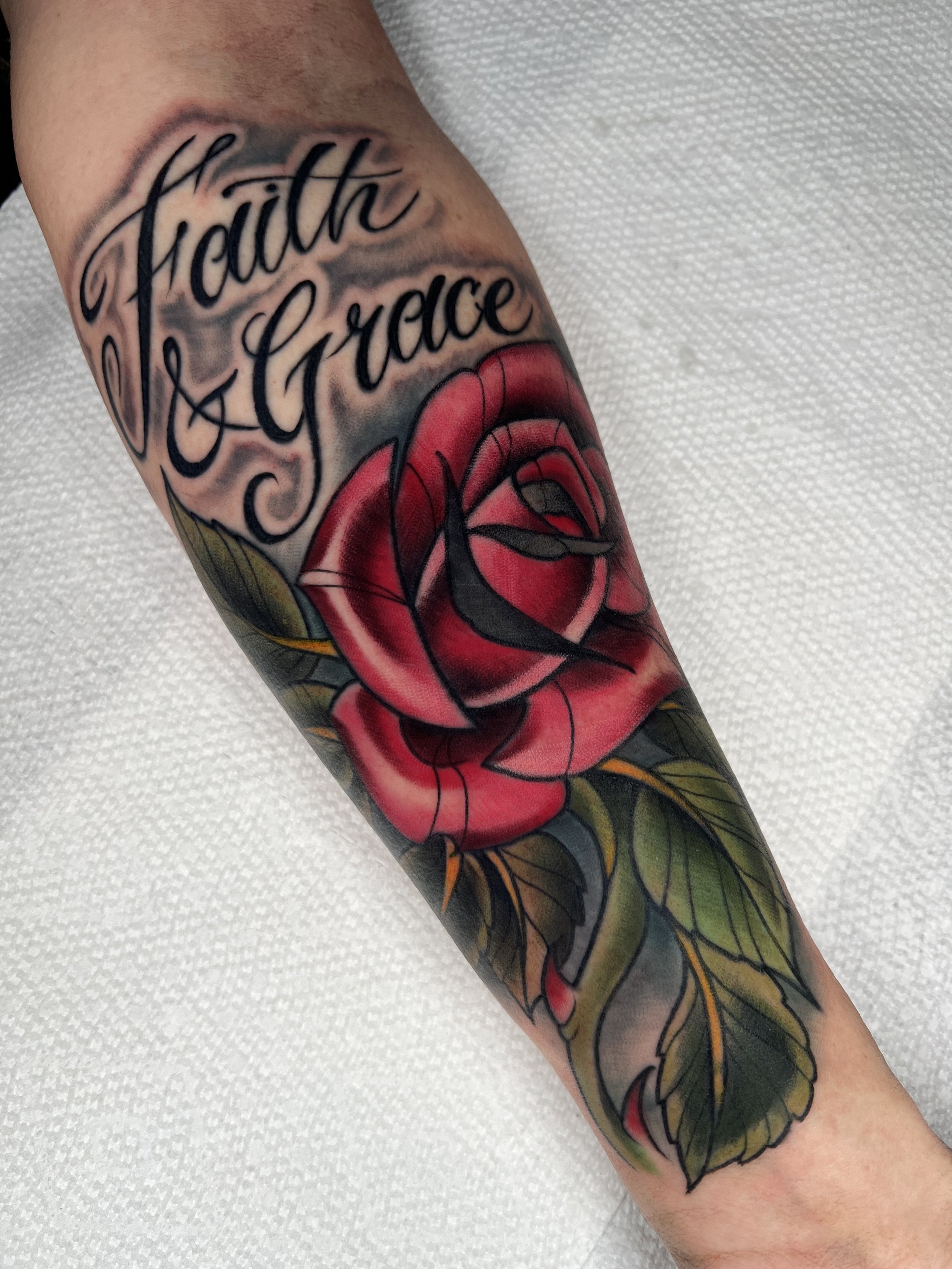 A tattoo on a person's arm with the words 'Faith & Grace' written in cursive, paired with a vibrant red rose and green leaves.