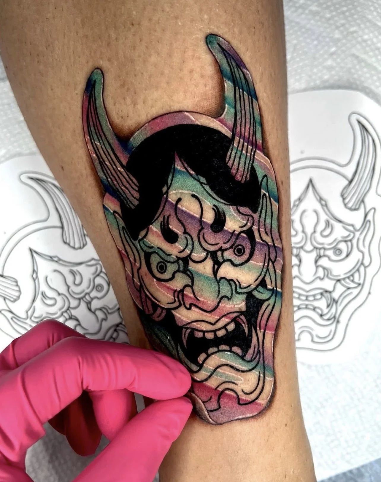 A colorful, stylized tattoo of a roaring demon or monster with exaggerated facial features and long, curved horns being inked onto a person's arm. The tattoo features bold outlines and vibrant, rainbow-like colors.