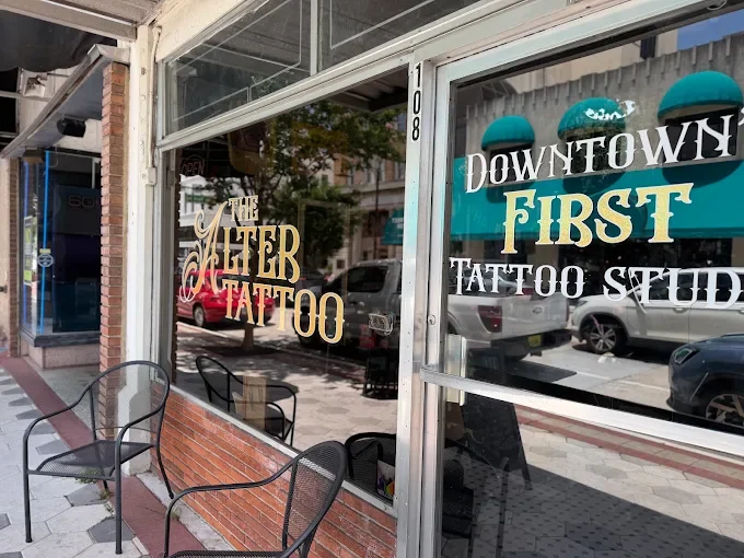 The storefront windows of a tattoo shop with signs advertising 'The Alter Tattoo' and 'Downtown First Tattoo Studio,' with outdoor seating including black chairs.