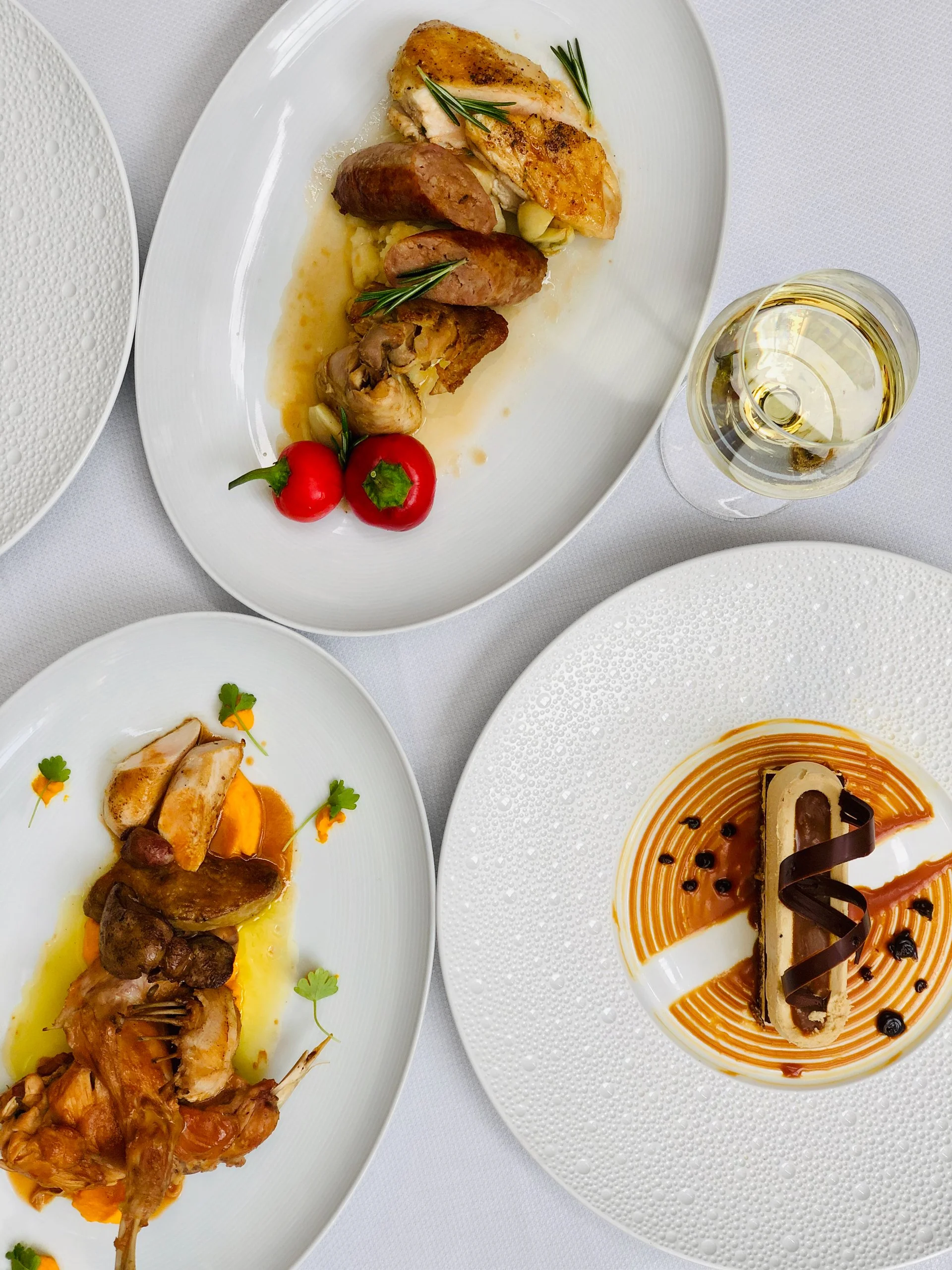 A plated meal with roasted chicken, sausage, and vegetables, a dish with seafood and vegetables, and a dessert with caramel sauce, chocolate, and berries on white plates, alongside a glass of white wine on a white tablecloth.