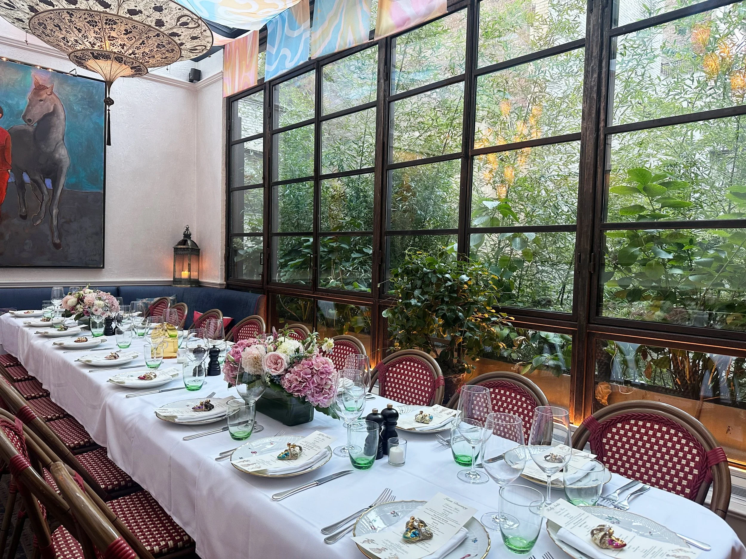 A long dining table set for a formal event with a white tablecloth, floral centerpieces, and place settings including glasses and silverware. The room has large windows with greenery outside, colorful artwork on the wall, and decorative umbrellas han
