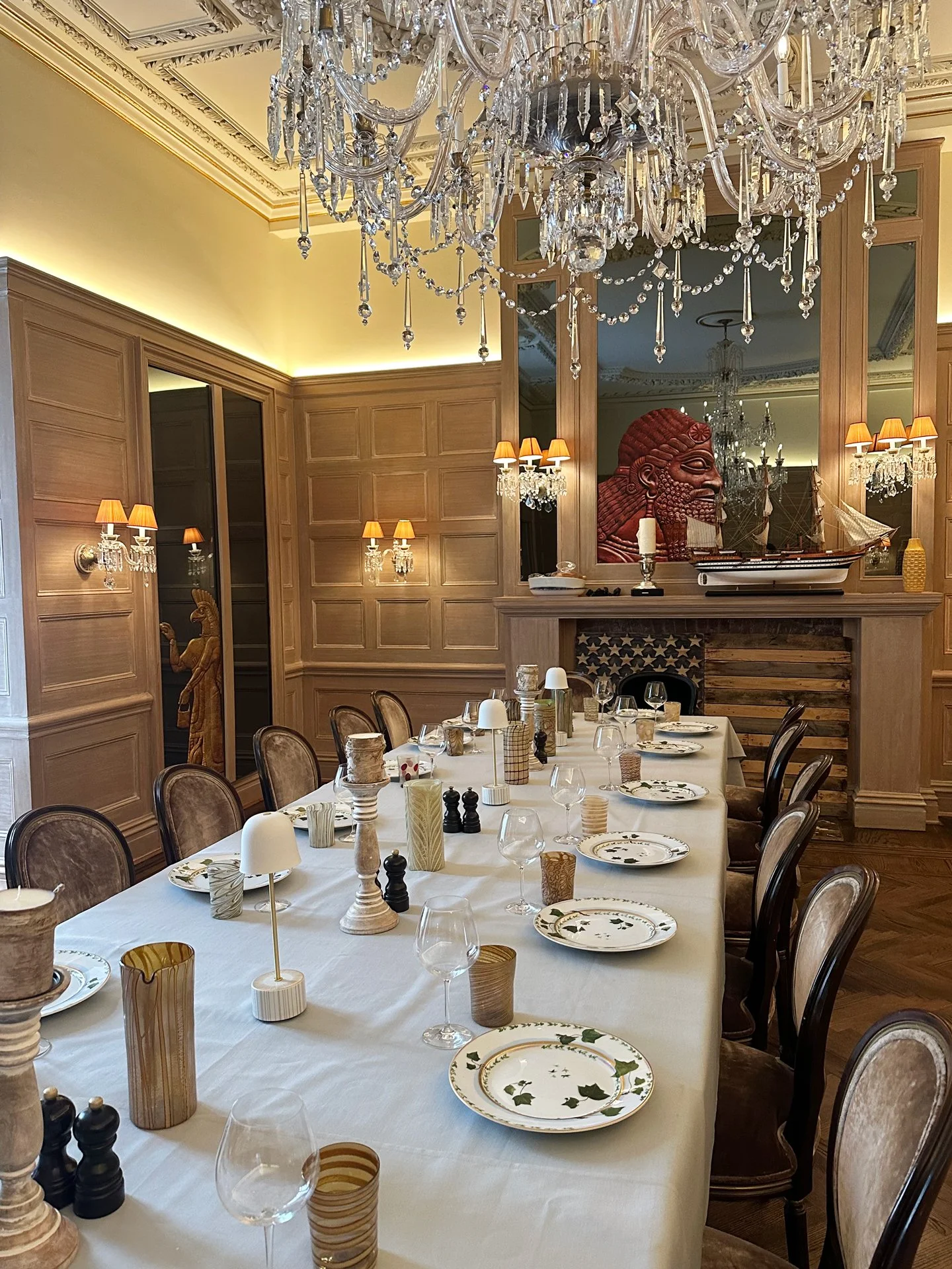 Elegant dining room with a long table set for a meal, surrounded by chairs, featuring a chandelier, wall sconces, wood-paneled walls, and decorative items including candles, vases, and a model ship. A large wooden sculpture of a face is reflected in 