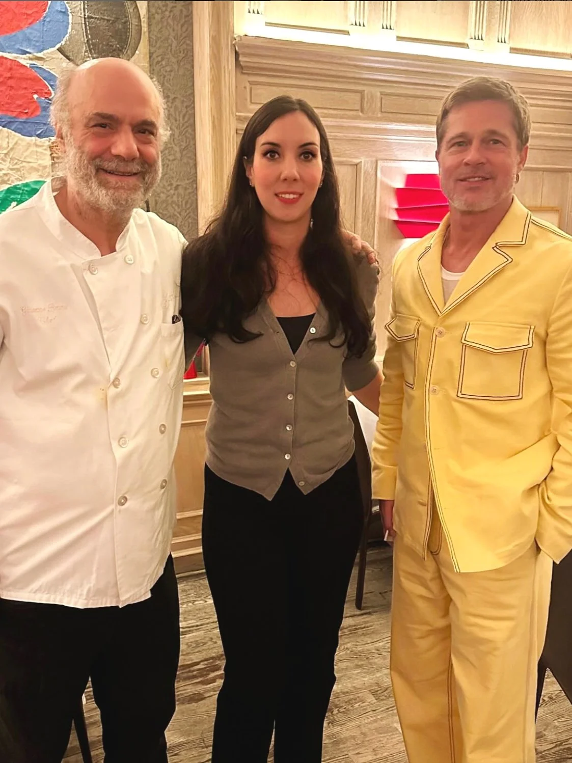 Three people standing together indoors with wooden wall paneling in the background. The person on the left is a man with a beard, wearing a white chef's coat. The person in the middle is a woman with long dark hair, wearing a gray button-up shirt and