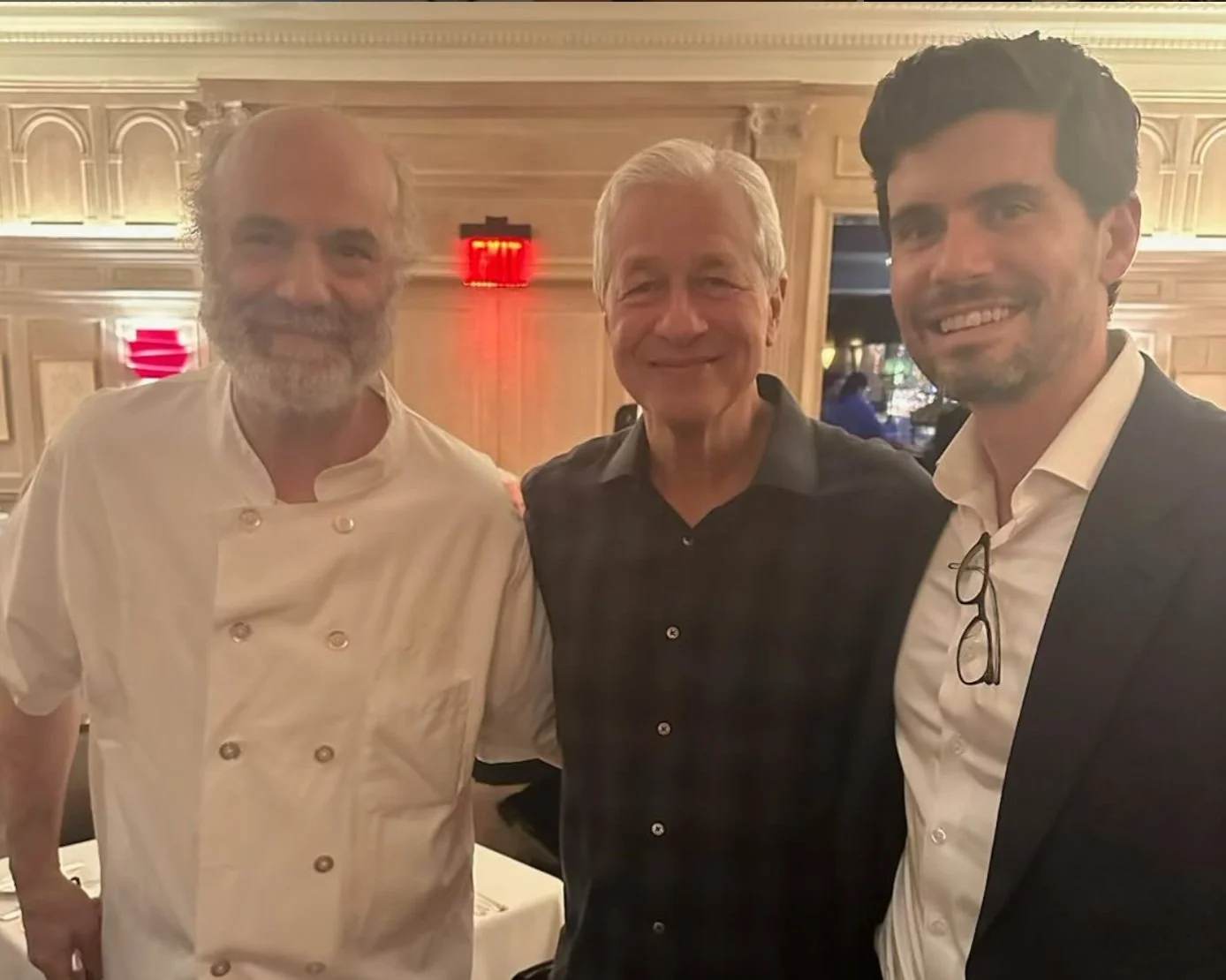 Three men smiling at a dinner event, standing close together inside a warmly lit, wood-paneled room.