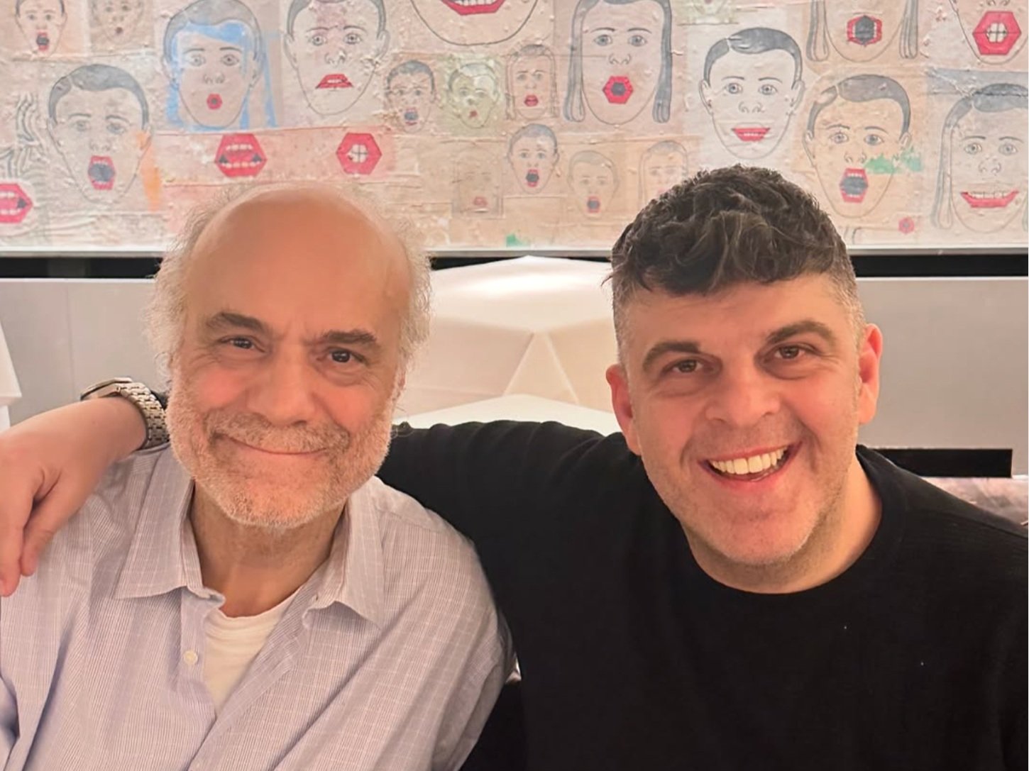 Two smiling men sitting with their arms around each other in a restaurant or cafe, behind them a colorful wall with illustrations of faces and some hexagon shapes.
