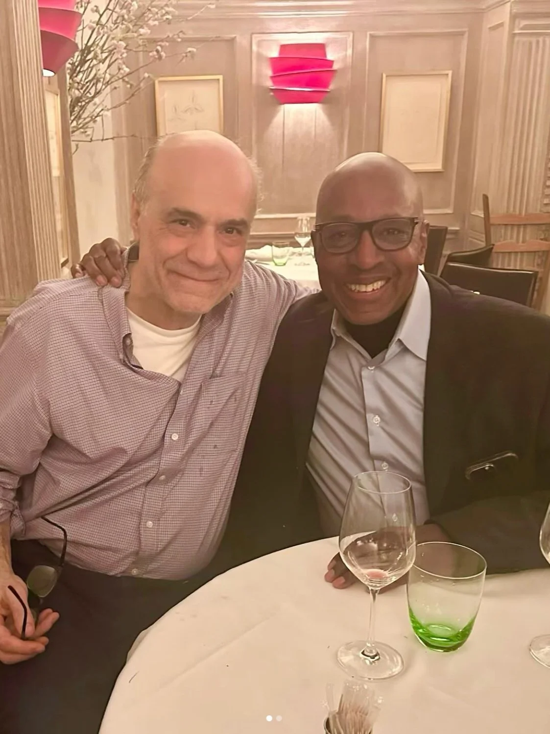 Two men sitting at a dinner table in a restaurant, smiling at the camera. One man has a light shirt, and the other wears glasses and a dark blazer. There are wine glasses and a green-tinted glass on the table. The background features wood-paneled wal