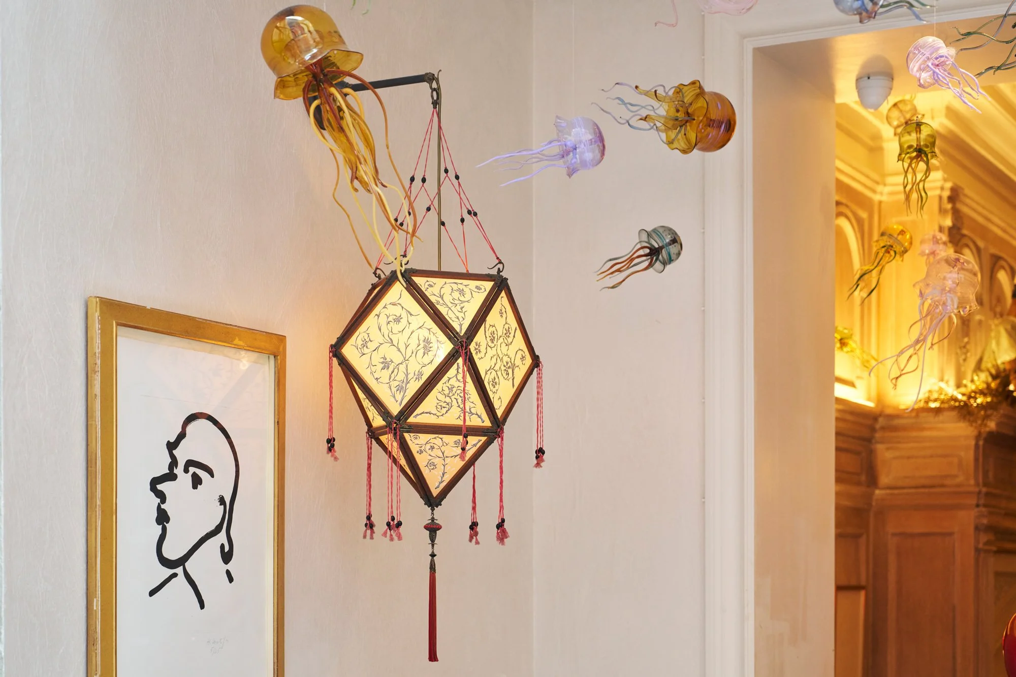 Decorative hanging lamp with intricate floral design, surrounded by hanging glass jellyfish-shaped ornaments in various colors, and a minimalist black and white line drawing of a female face on the wall in a well-lit interior space.
