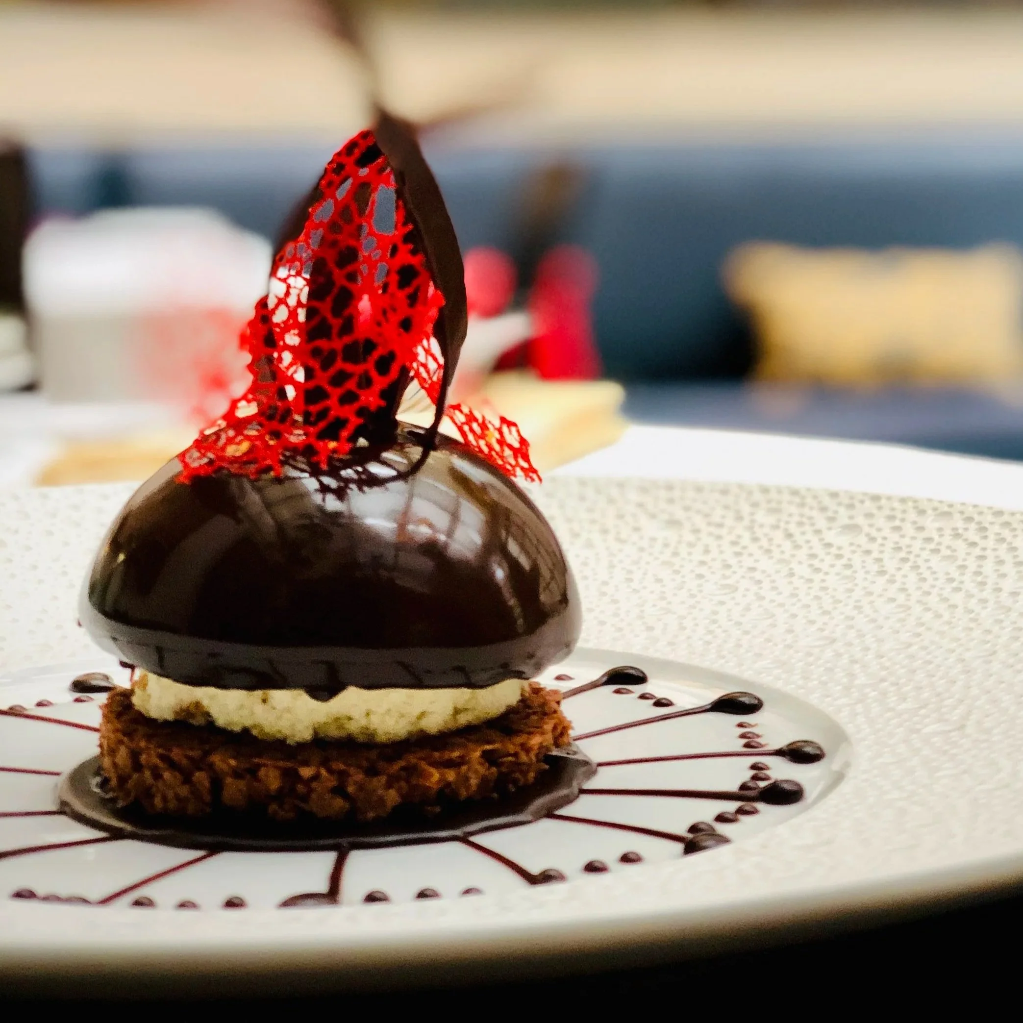 A gourmet chocolate dessert with a shiny mirror glaze, topped with a decorative red chocolate lattice and a chocolate strip, served on a white plate with a chocolate sauce spiral design.