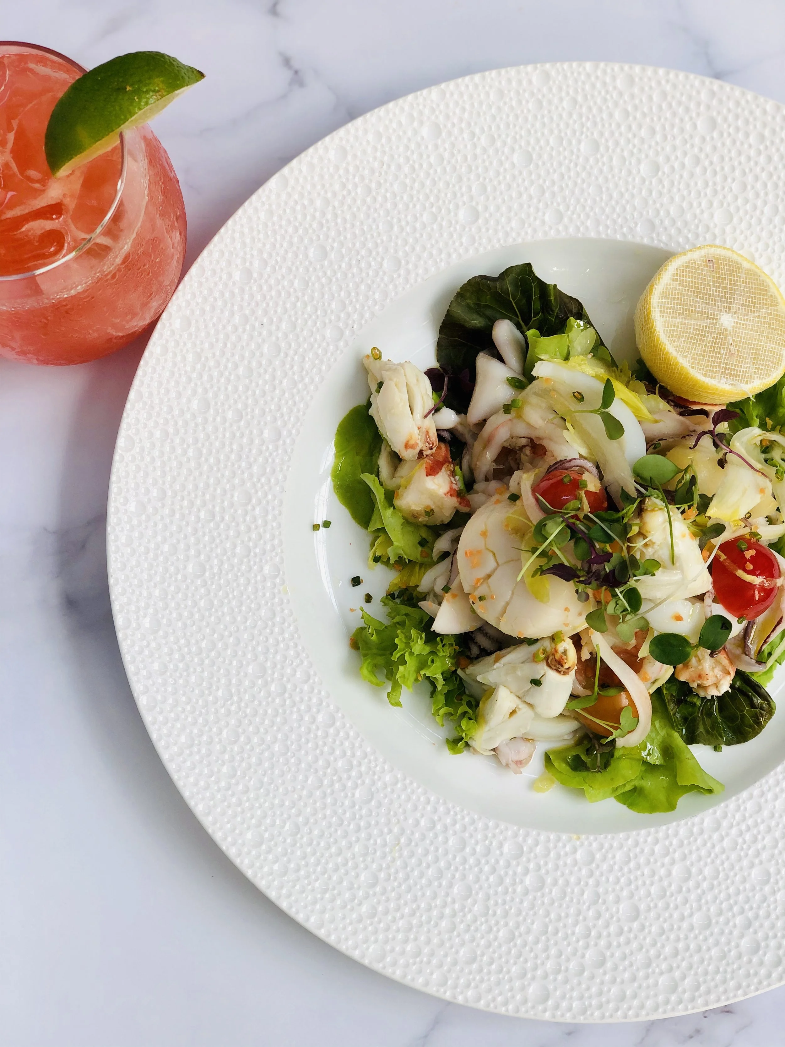 A seafood salad with lettuce, cherry tomatoes, and microgreens on a white textured plate, with a halved lemon on the side, and a pink beverage with a lime wedge in a glass to the side.