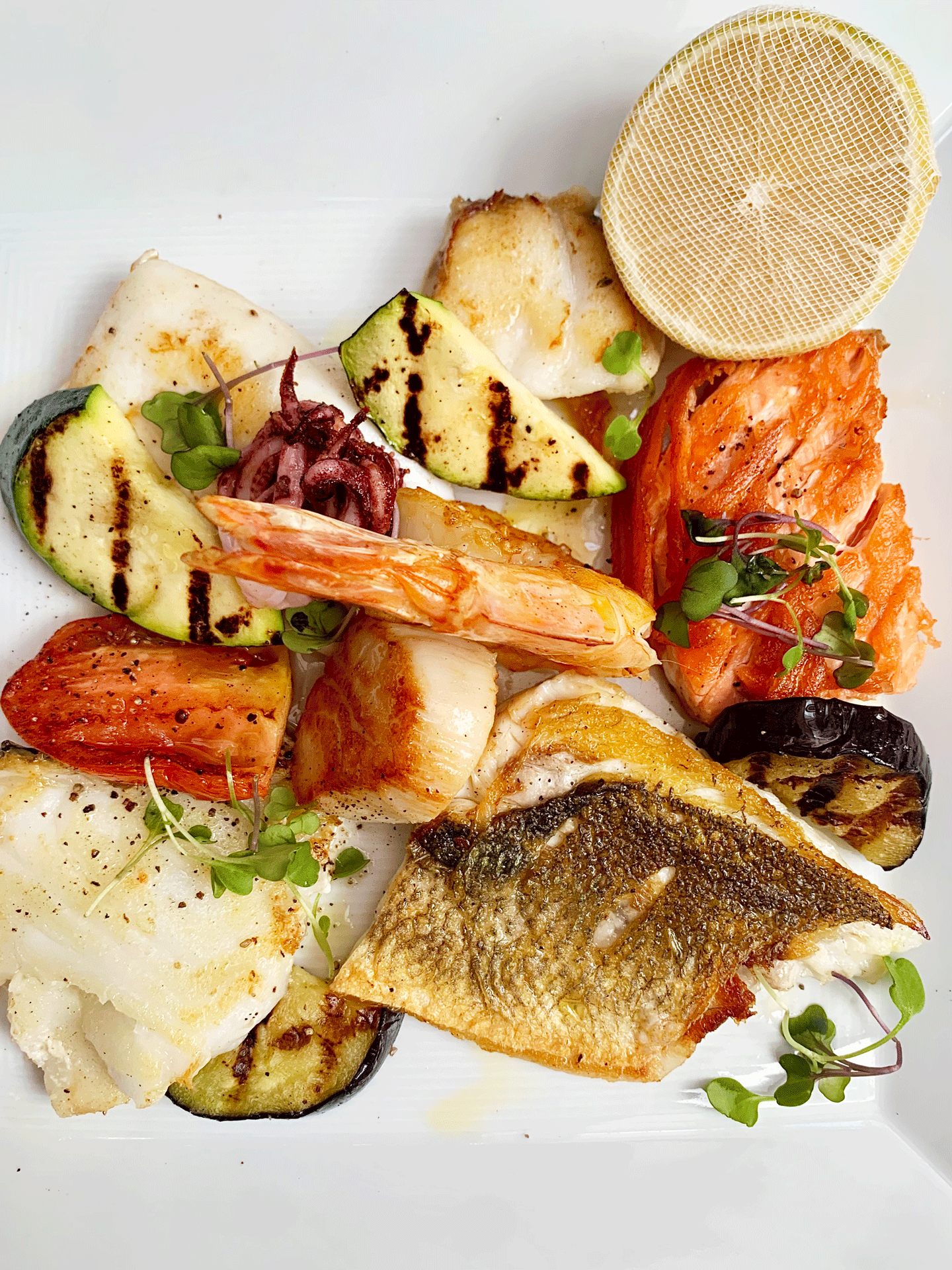 Assorted grilled seafood, vegetables, and microgreens on a white plate, including fish, shrimp, zucchini, tomato, and lemon, with grilled lemon slices and a round netted lemon.