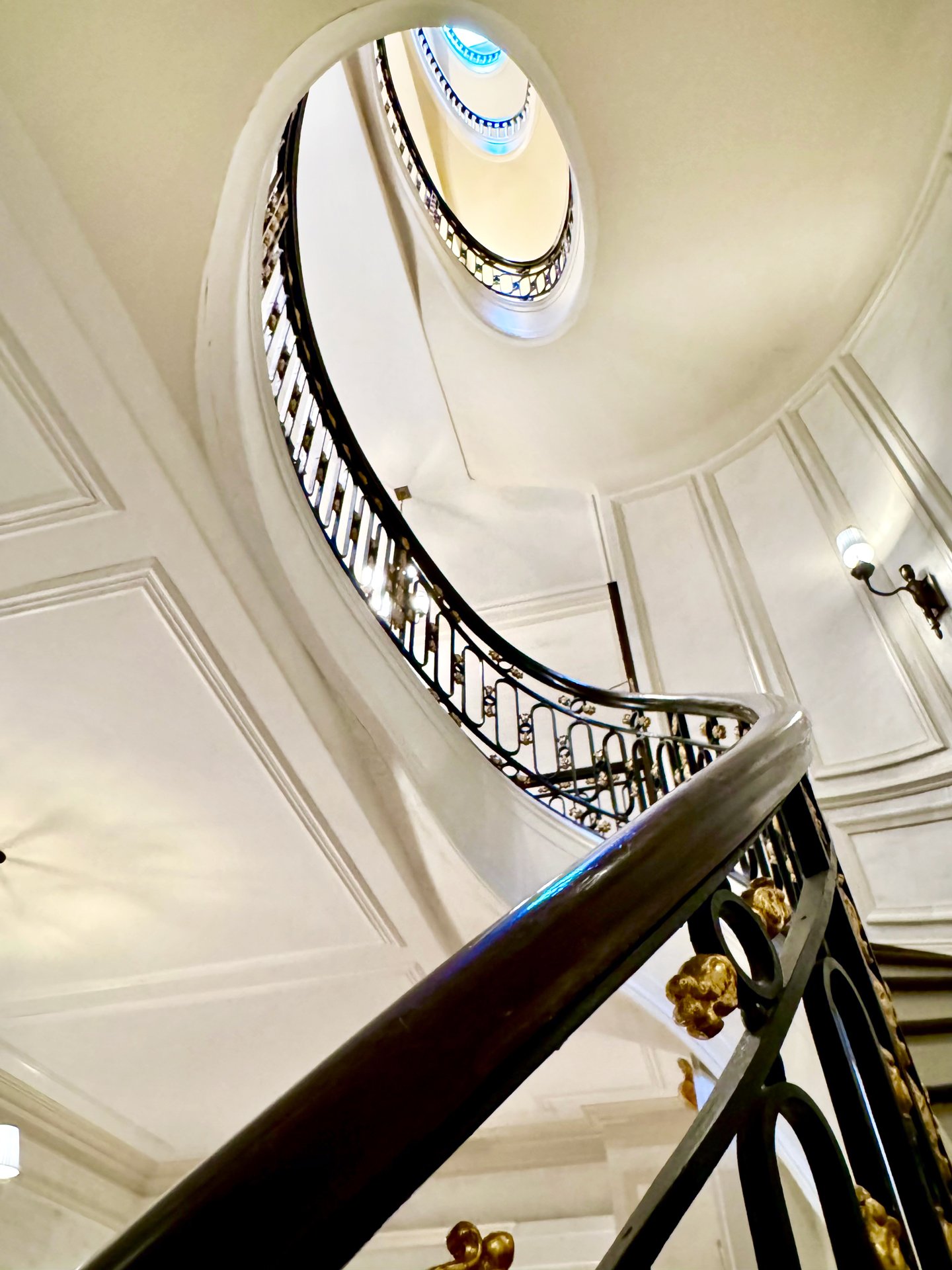 A spiral staircase viewed from bottom to top, with black and gold ornate railing, white walls, and a ceiling with decorative moldings and wall-mounted light fixtures.