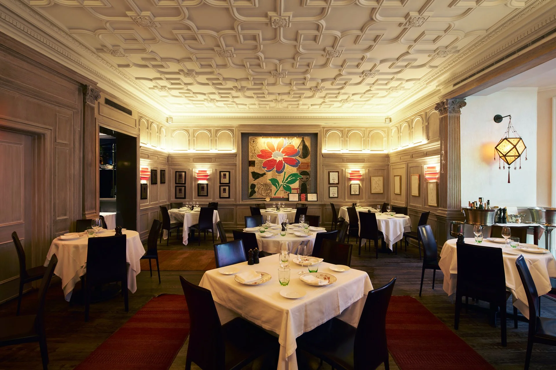 Elegant restaurant dining room with white-draped tables, black chairs, soft warm lighting, a large colorful floral painting on the back wall, and decorative wall panels.