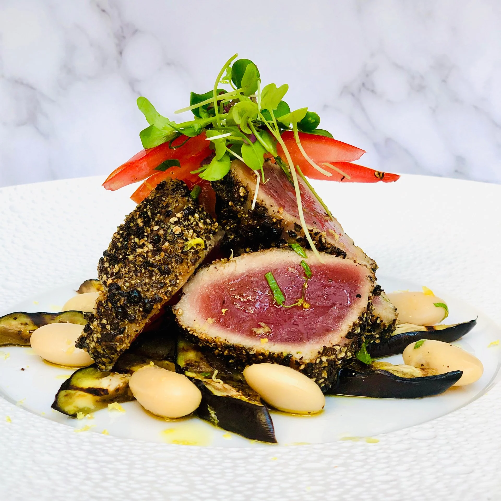 Seared tuna steak coated with cracked black pepper and herbs, served with white beans, grilled eggplant, and garnished with tomato slices, microgreens, and lemon zest