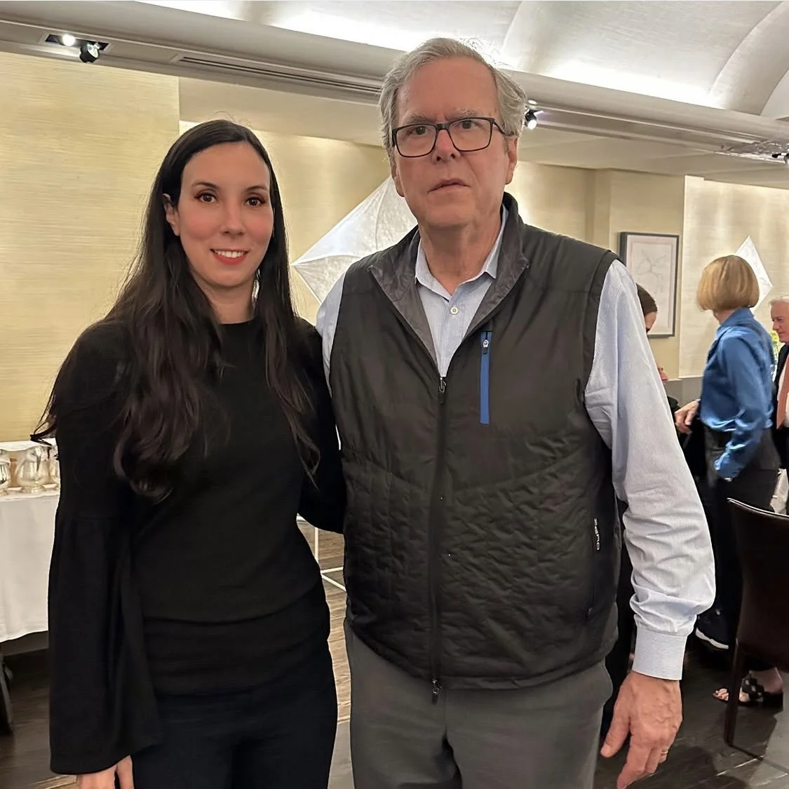 A young woman and an older man are standing together indoors, posing for the photo. The woman has long dark hair and is wearing a black top, smiling. The man has gray hair, glasses, and is wearing a light blue shirt with a black vest, looking serious