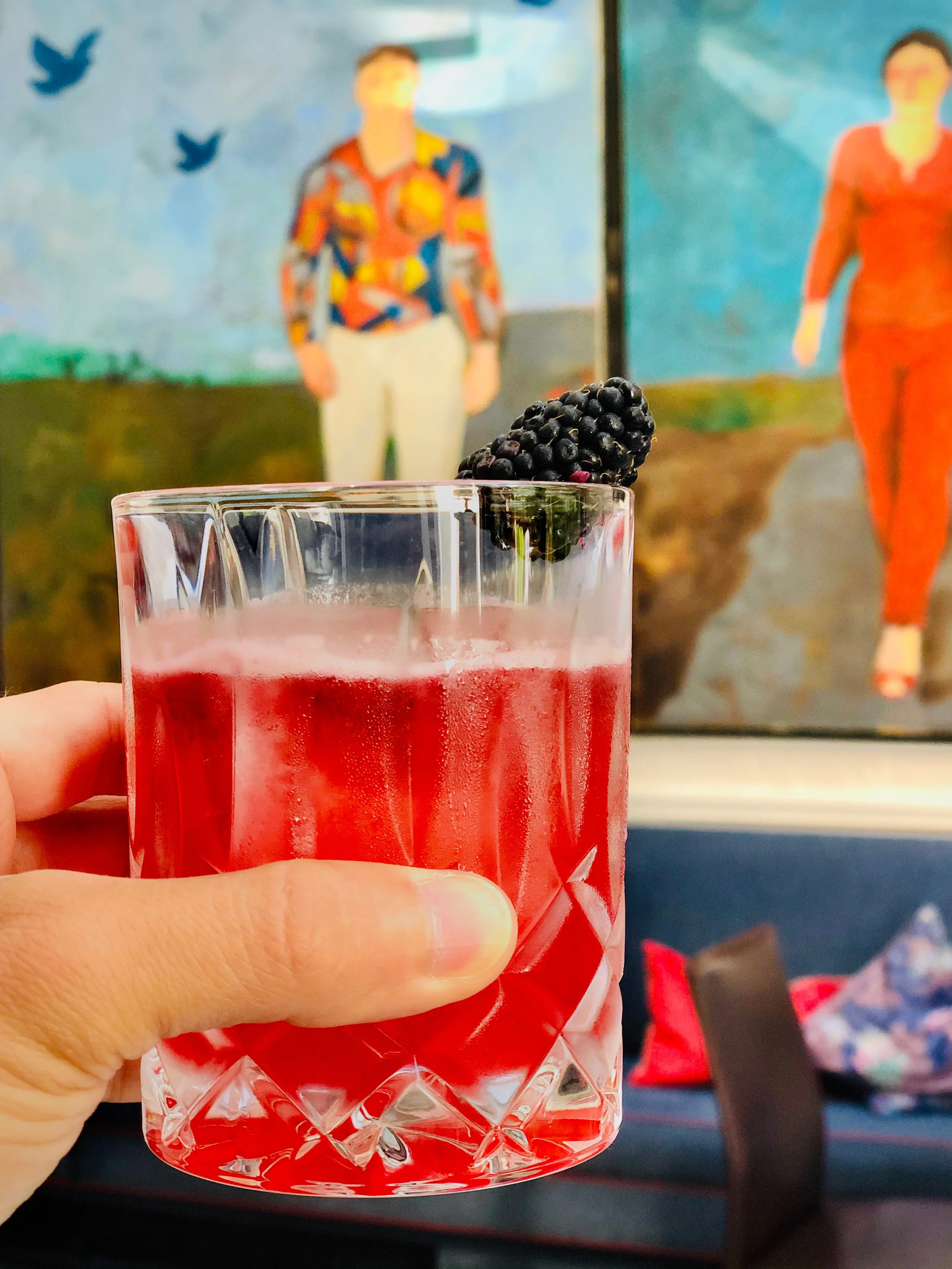 A hand holding a glass of red beverage garnished with a blackberry. In the background, there is a painting of two women walking and birds flying in the sky.