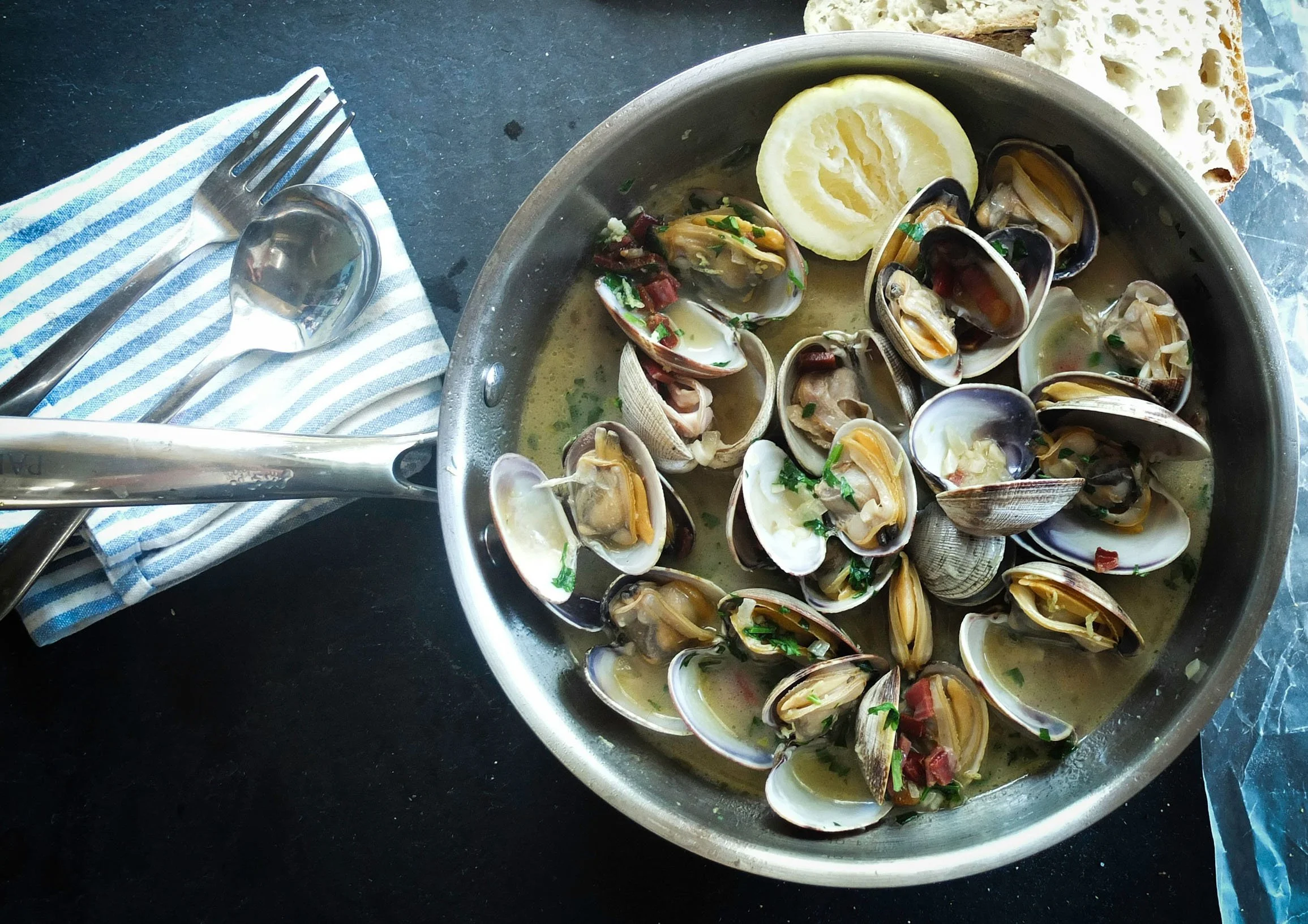 Pan of cooked clams in broth garnished with chopped herbs, served with lemon slices and bread on the side.