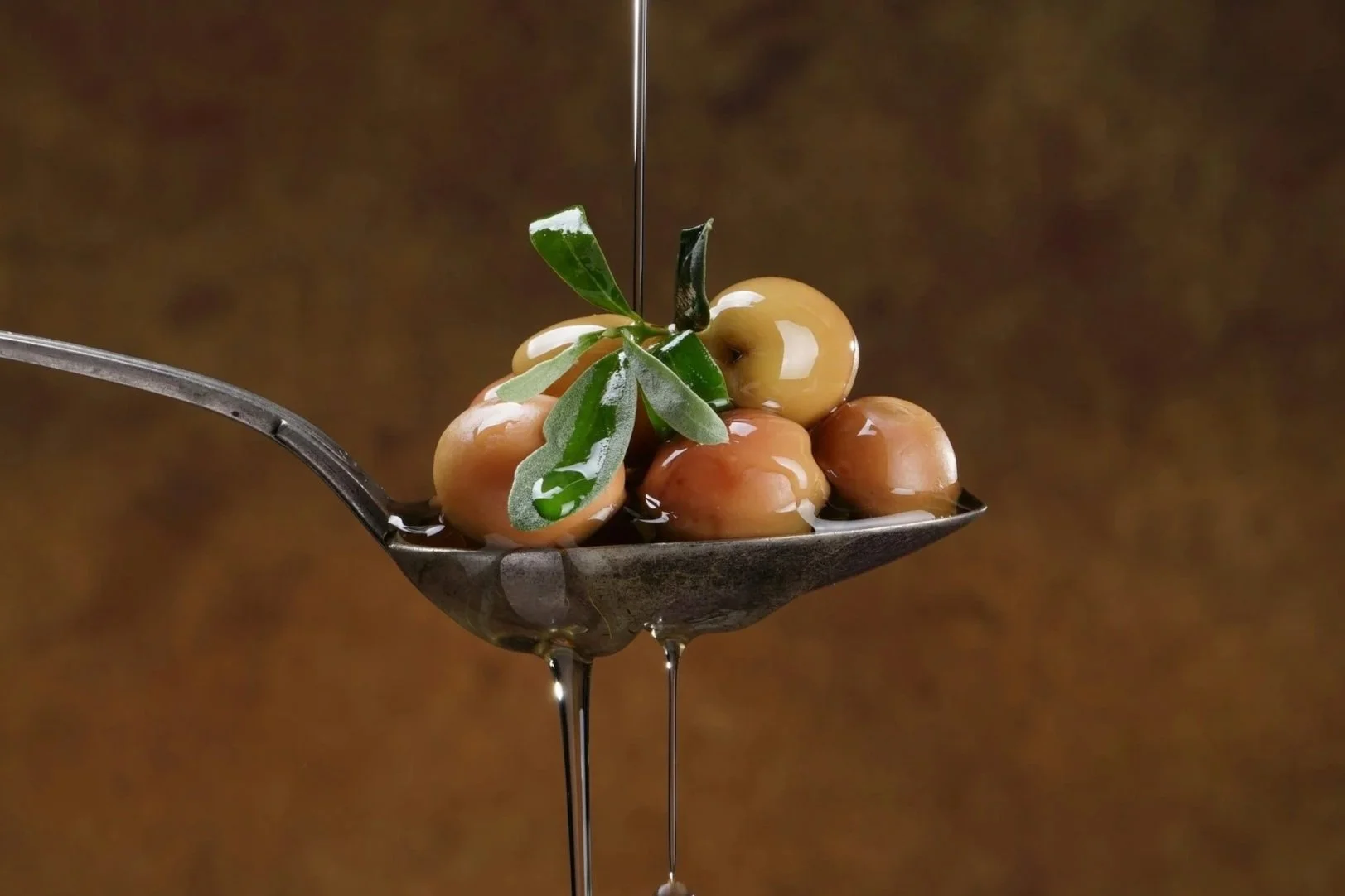 A silver spoon holding several green and brown olives, topped with a sprig of green olive leaves, with oil being drizzled over them.