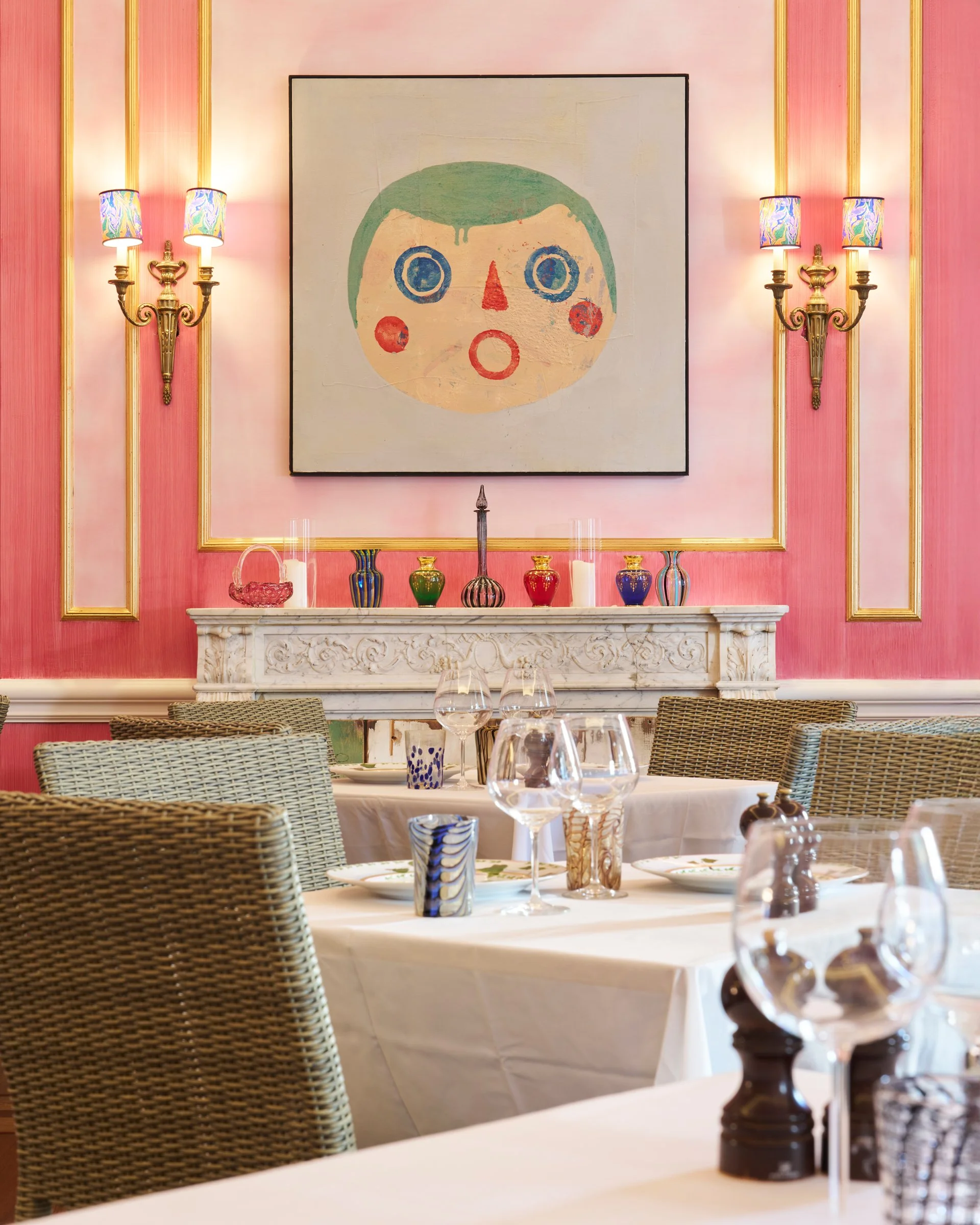 A pink dining room with a white marble fireplace and colorful vases, a large abstract face painting, and tables set with glassware and small salt and pepper shakers.