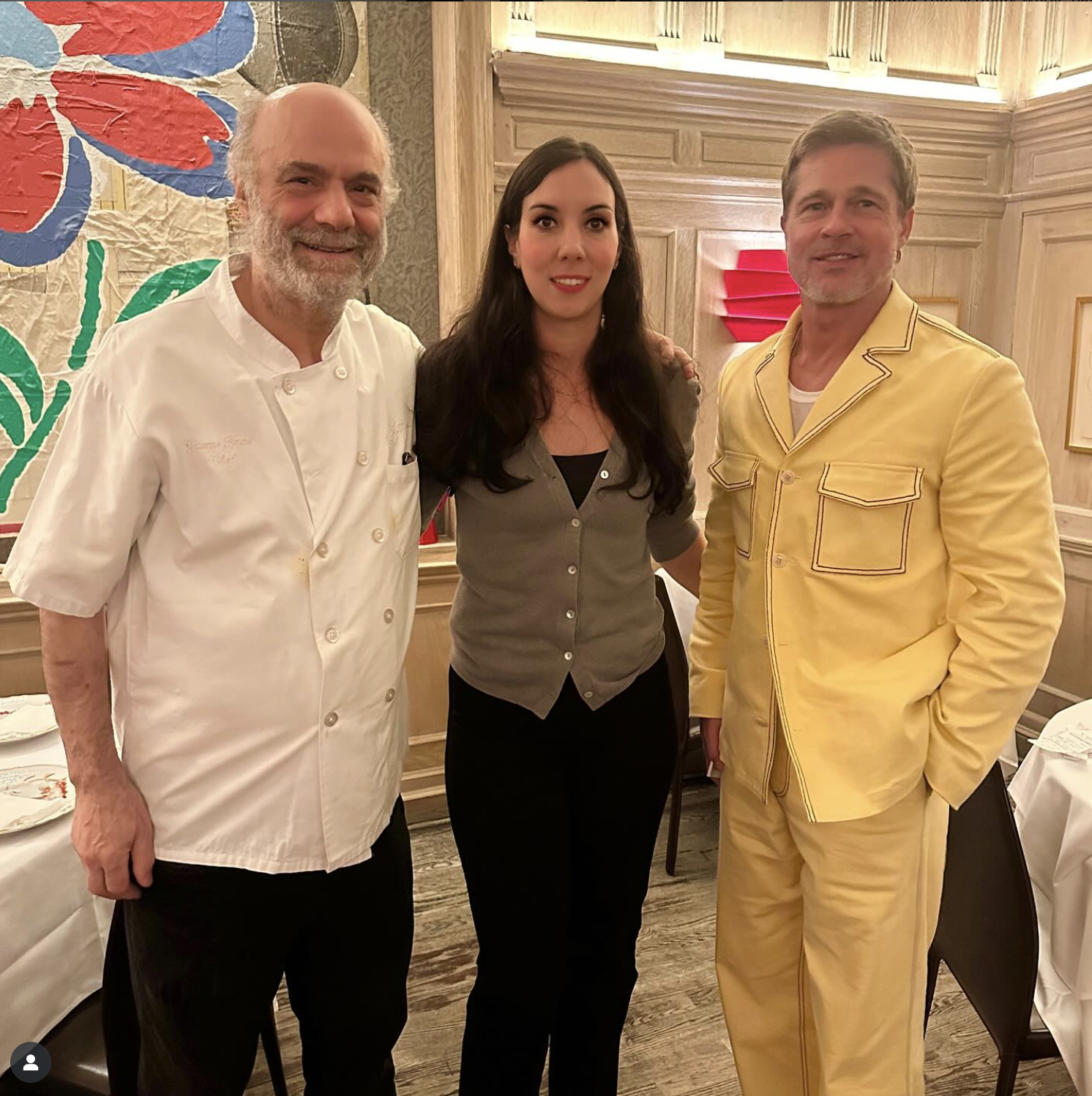 Three people standing together in a restaurant or dining area, smiling at the camera. The person on the left is a chef wearing a white chef's coat, the woman in the middle has long dark hair and is dressed in a gray button-up shirt, and the person on