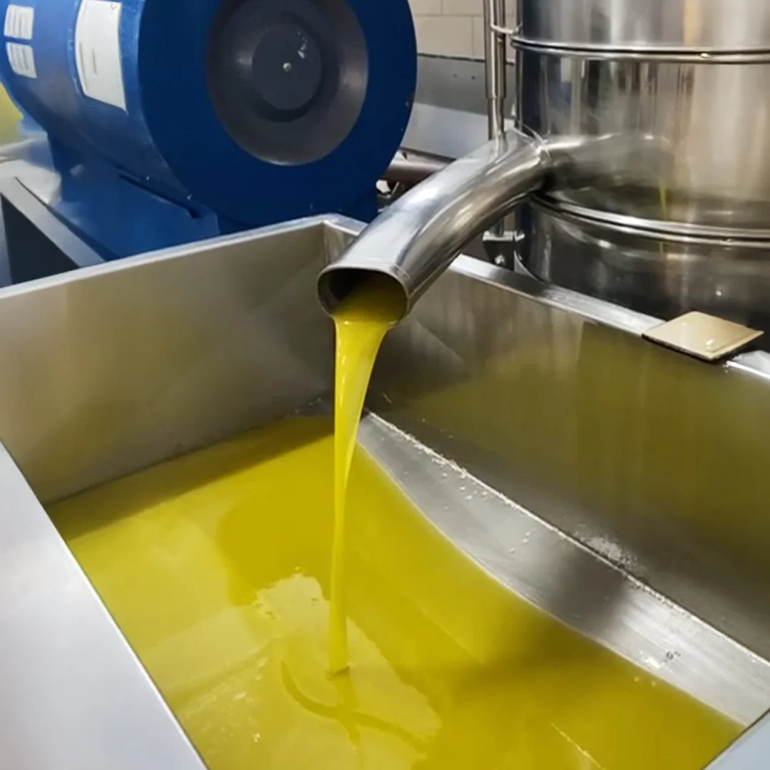 Yellow liquid flowing from a metal pipe into a large stainless steel container in an industrial setting.