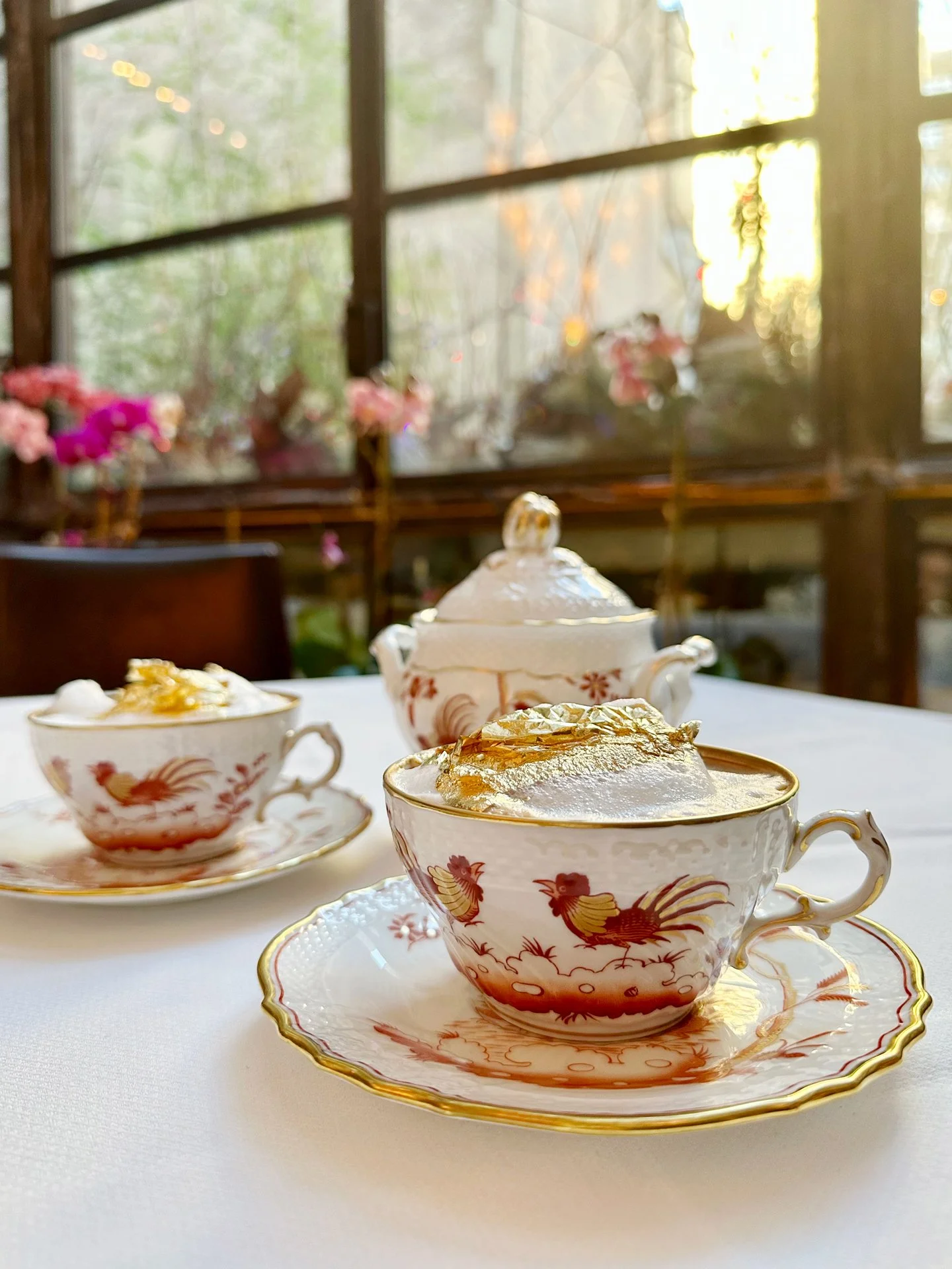 Tea cups with gold accents and rooster design, filled with tea and topped with gold leaf, on a white tablecloth in a sunlit room with large windows and pink flowers in the background.