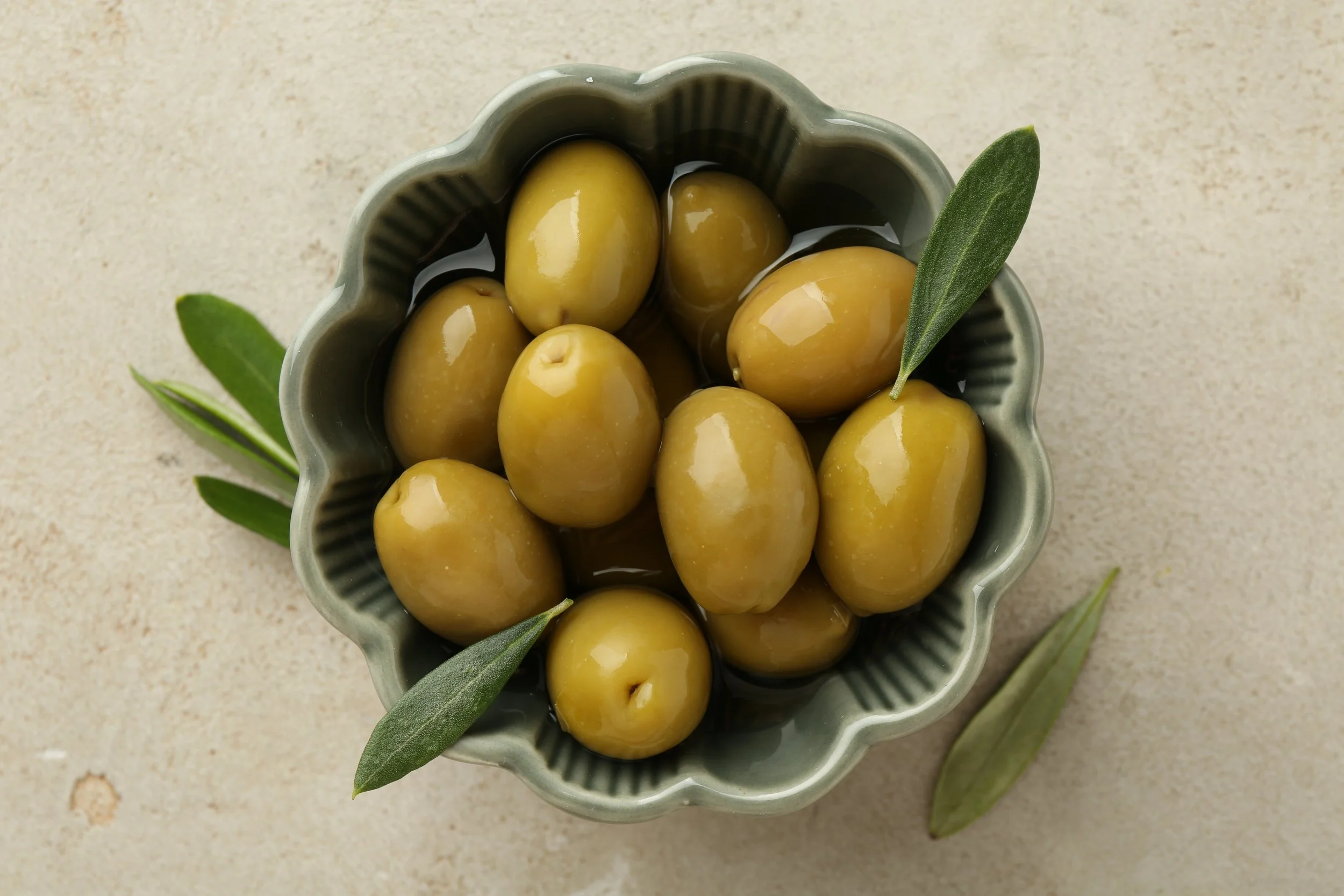 A bowl of green olives with olive leaves on a light-colored surface.