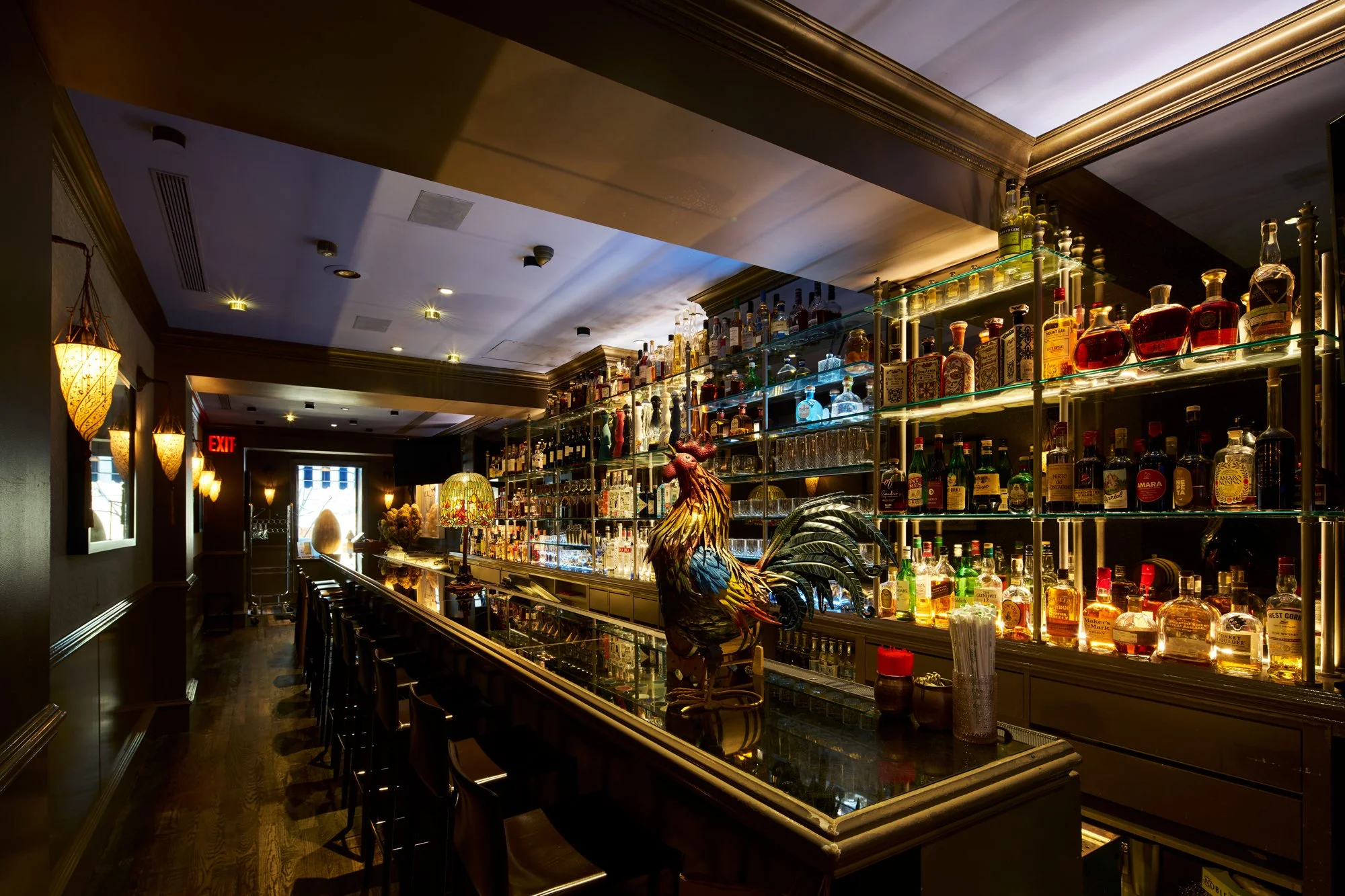 An elegant bar interior with a long counter, shelves filled with bottles of liquor, a colorful rooster sculpture on the counter, and warm lighting.