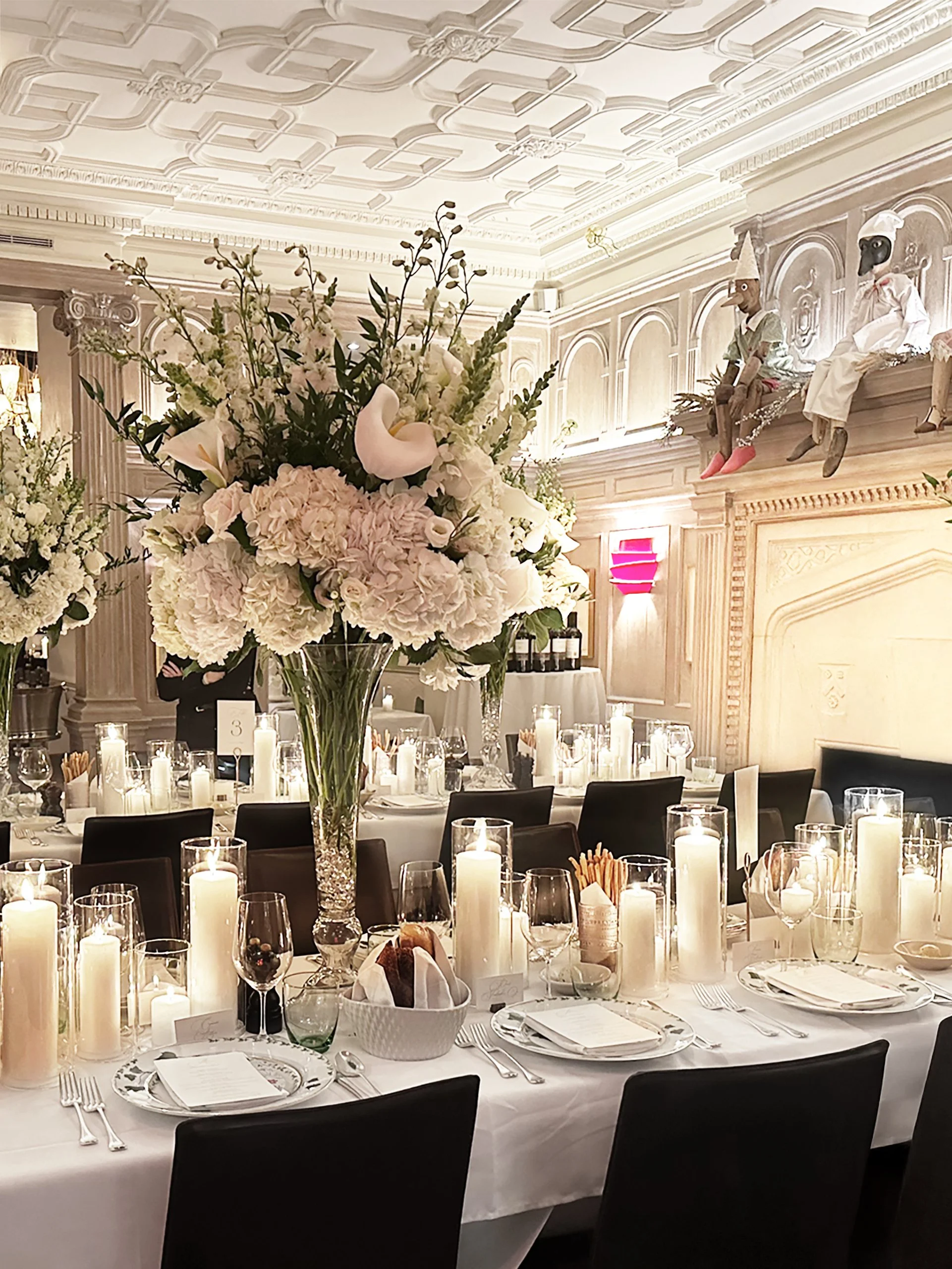 Elegant banquet table decorated with a large floral arrangement, candles, and place settings in a lavish room with ornate ceiling and wall decor, featuring two dolls dressed as chefs sitting on a ledge.