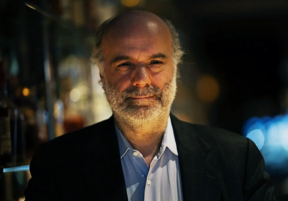 Portrait of an older man with gray hair and a beard, wearing a dark suit jacket and white shirt, indoors with blurred lights in the background.