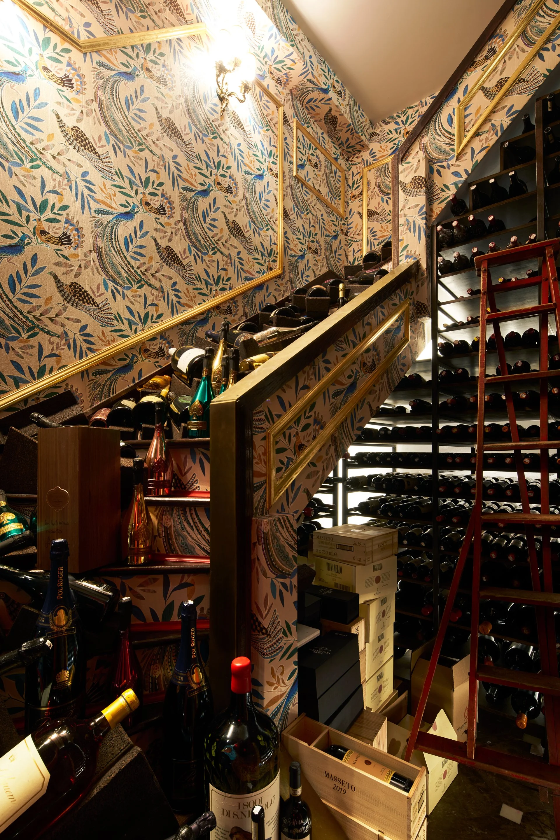 A wine cellar with bottles stored on shelves and stacked boxes, decorated with patterned wallpaper featuring peacocks and leaves, and illuminated by a wall-mounted light fixture.