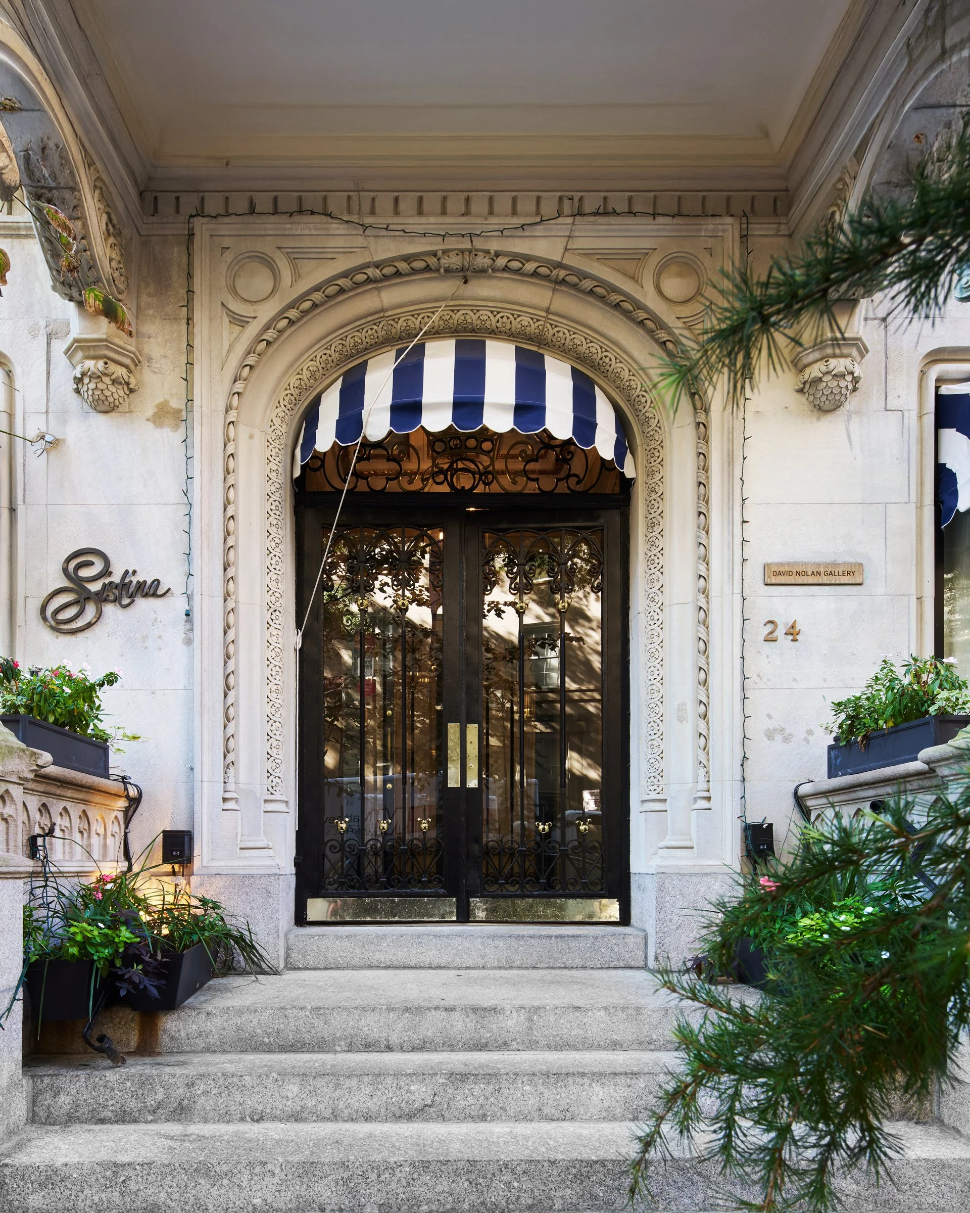 Concrete front steps leading up to a decorative black metal gate with intricate designs, framed by an ornate stone archway with a blue and white striped awning overhead, flanked by flower pots with green plants and flowers, and a sign reading 'Sestin