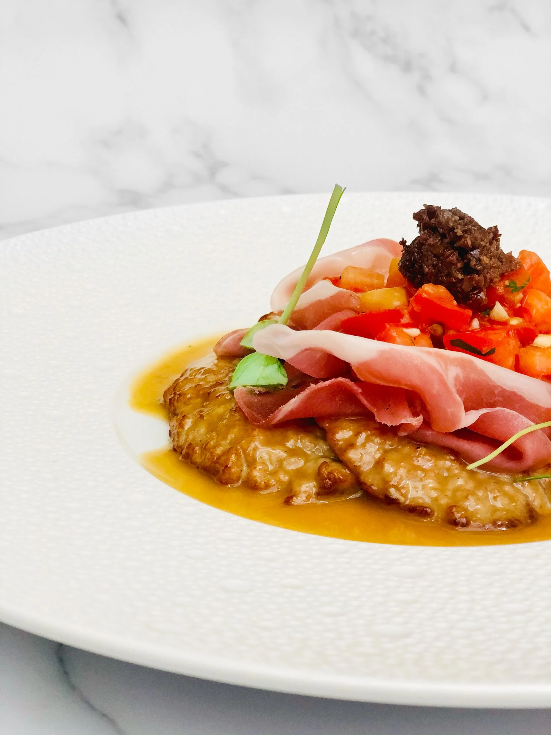 A plated gourmet dish with a piece of meat topped with a sauce, garnished with greens, alongside sliced ham, chopped vegetables, and a dollop of dark tapenade on a white textured plate.