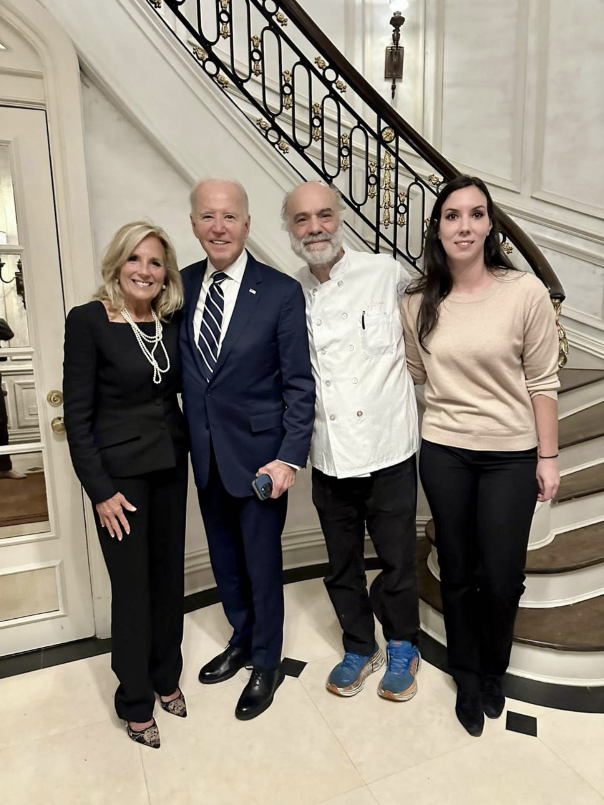 Four people standing in front of a staircase, smiling for a photo. One woman in a black outfit with pearl necklace, one man in a suit and striped tie, one man in a chef's coat and sneakers, and one woman in a beige sweater and black pants.